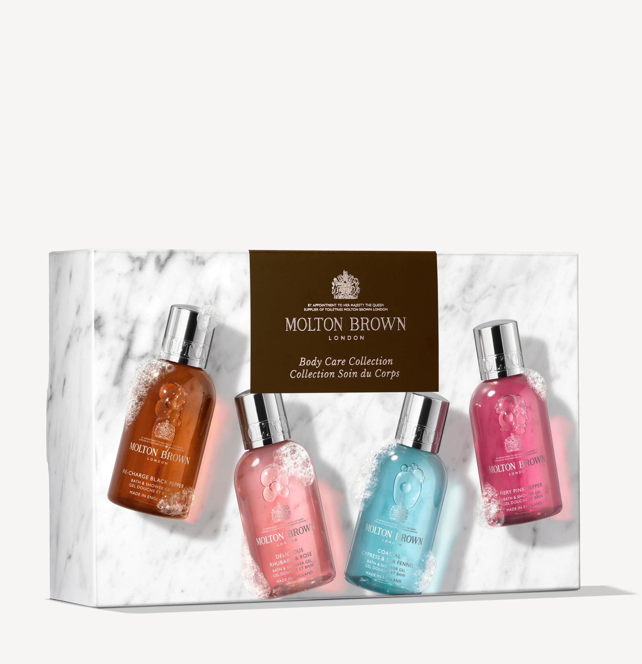 The MoltonBrown Nederlands Woody & Floral Body Verzorgingscollectie features four douchegel bottles in amber, pink, blue, and magenta, arranged in a white marble box with a gold label.