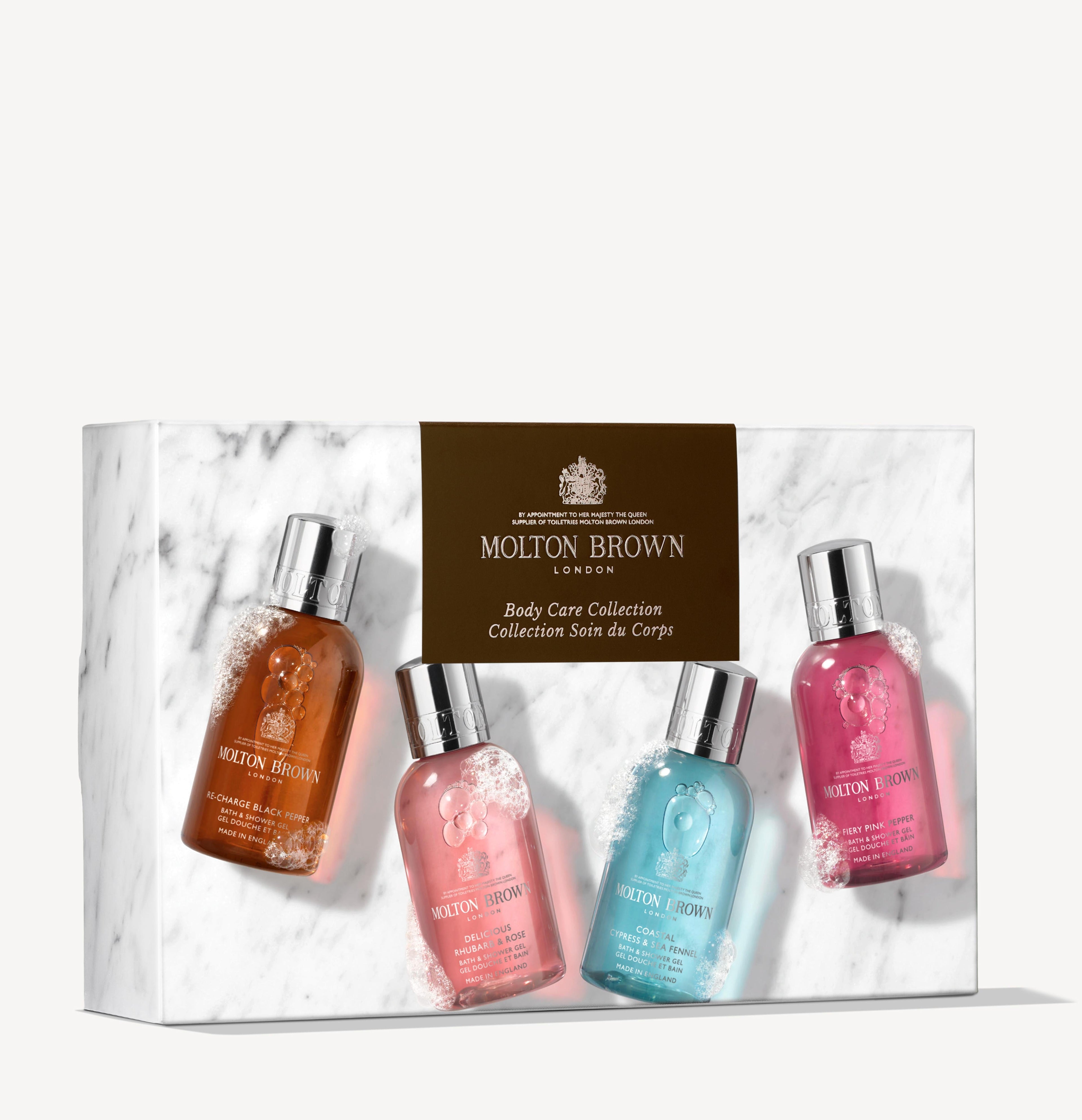 The MoltonBrown Nederlands Woody & Floral Body Verzorgingscollectie features four douchegel bottles in amber, pink, blue, and magenta, arranged in a white marble box with a gold label.