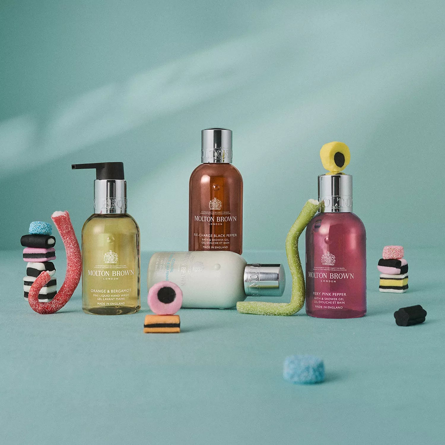 Four MoltonBrown Nederlands Fresh & Floral Hand Verzorgingscollectie products are arranged on a light blue surface with colorful candy and licorice, highlighting a luxurious hand care experience against a soft teal gradient background.