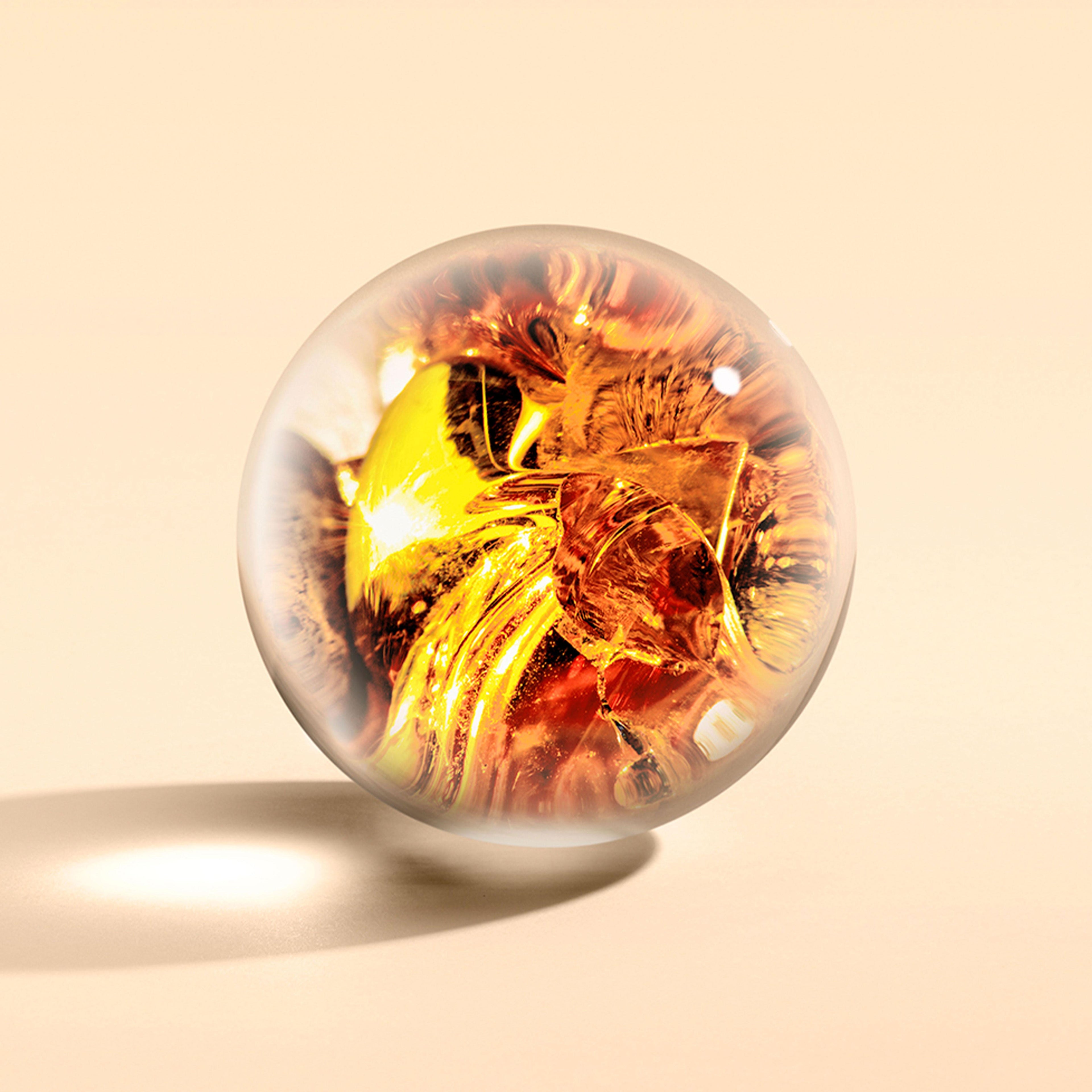 The Neon Amber Bad- & Douchegel 300ml by MoltonBrown Nederlands, with its clear glass sphere and intricate amber-gold swirls, rests on a smooth beige surface, casting a soft shadow.
