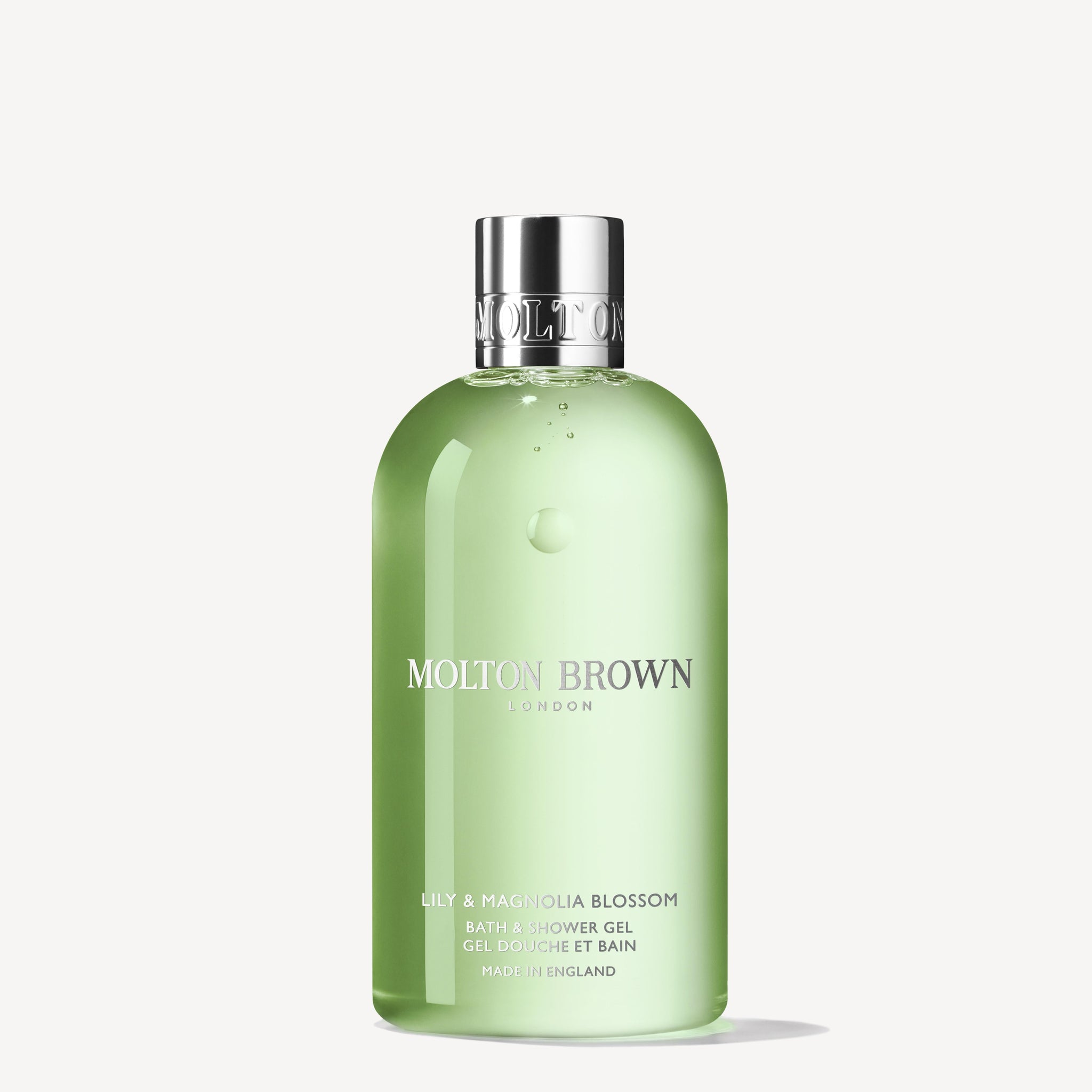A clear green bottle of MoltonBrown Nederlands Lily & Magnolia Blossom Bad- & Douchegel 300ml with a silver cap, een wit-bloemige douchegel geïmporteerd uit Engeland, shown against a plain white background.