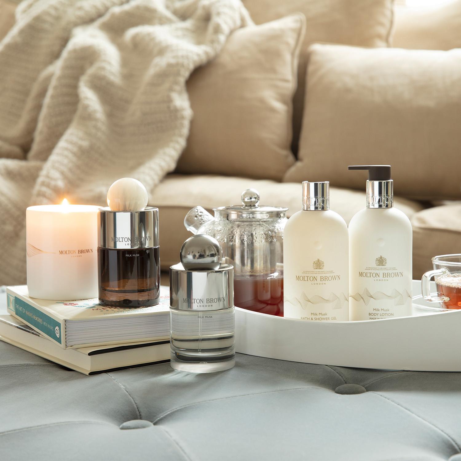 A cozy scene with MoltonBrown Nederlands Milk Musk Bodylotion 300ml to hydrate the skin, a lit candle, tea in a glass pot, two cups, and books on a tray atop a soft ottoman, with a beige couch and blanket in the background.