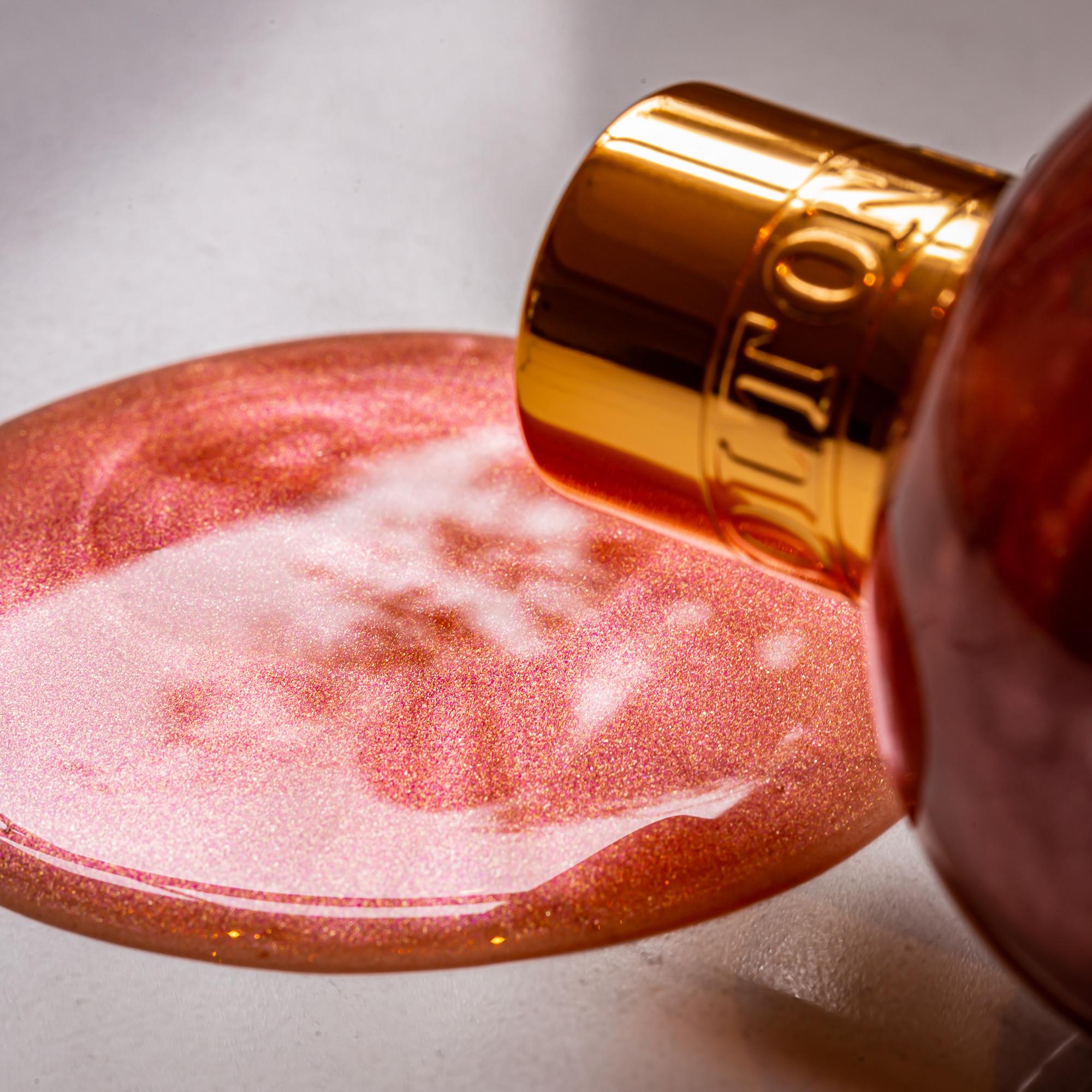 A close-up of a gold-capped MoltonBrown Nederlands Rose Dunes Bad- & Douchegel 300ml bottle spills shimmery pink gel onto a smooth surface, forming a reflective, glossy pool.