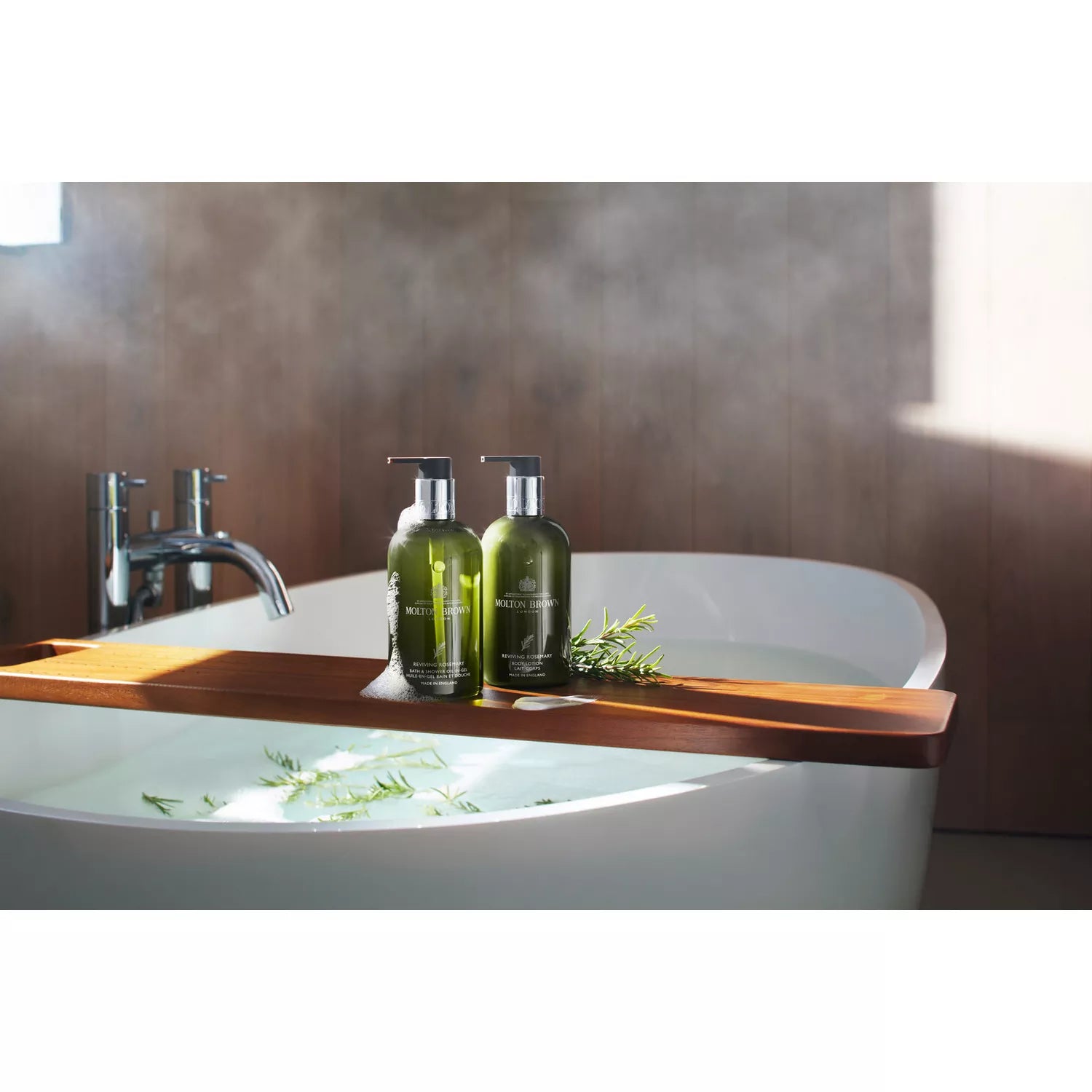 Two green pump bottles of Reviving Rosemary Bad- & Doucheolie-in-Gel by MoltonBrown Nederlands are on a wooden bath tray over a steaming white tub, with sprigs of rosemary eucalyptus nearby, sunlight adding to the spa-like, aromatic atmosphere.
