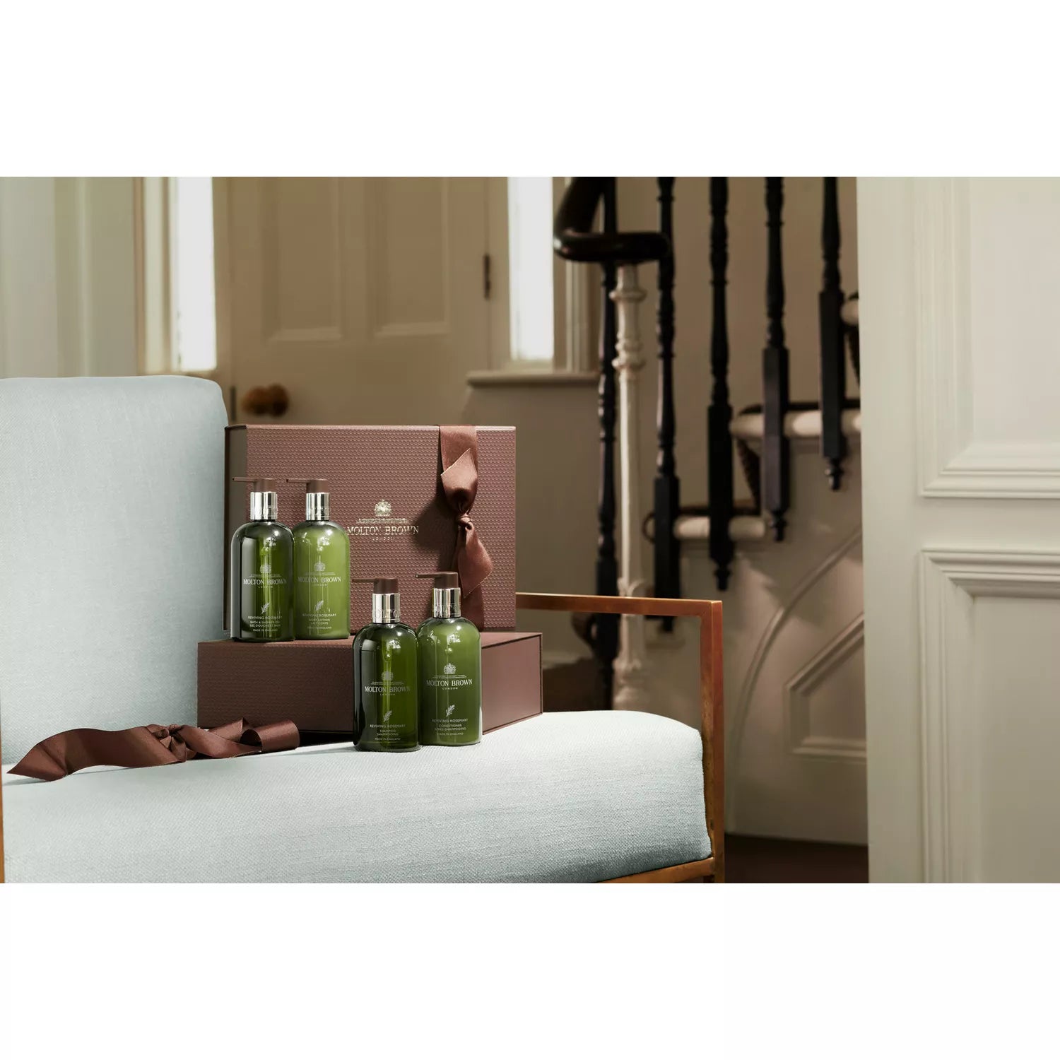 A gift set of four green bottles of MoltonBrown Nederlands Reviving Rosemary Bad- & Doucheolie-in-Gel in a brown ribboned box, displayed on a light grey chair with soft lighting, a staircase, and white door in the background.
