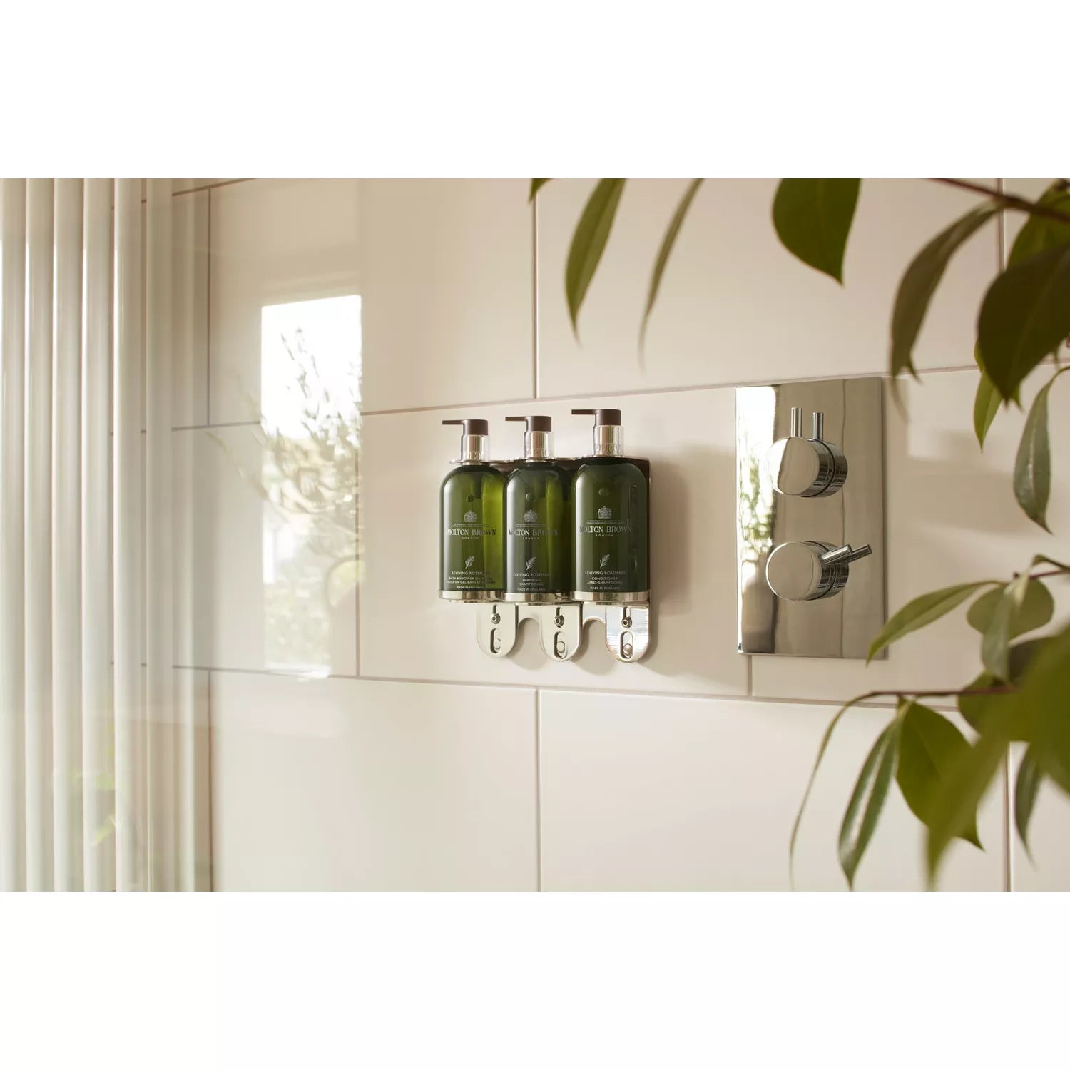 Three green pump bottles of MoltonBrown Nederlands Reviving Rosemary Bad- & Doucheolie-in-Gel are mounted on a tiled shower wall beside a shiny metal control panel, with leafy greenery catching sunlight through vertical blinds.