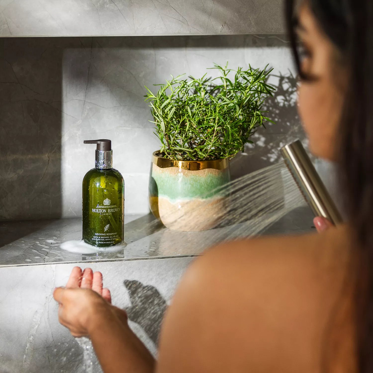 A person in a shower holds their hand under running water near a green bottle of MoltonBrown Nederlands Reviving Rosemary Bad- & Doucheolie-in-Gel and a potted rosemary eucalyptus plant on a glass shelf.