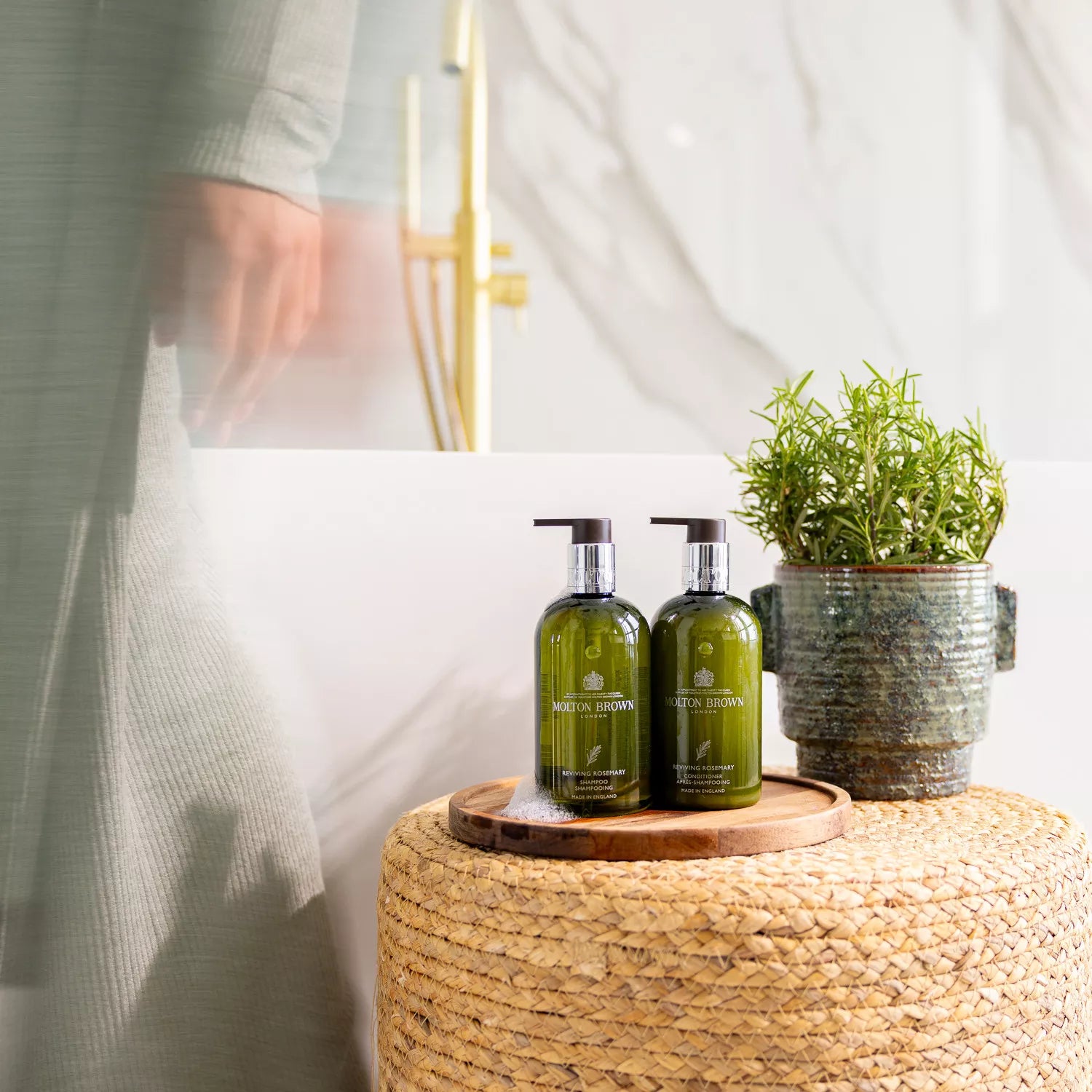 Two green bottles of MoltonBrown Nederlands, including Reviving Rosemary Shampoo, sit on a wooden tray atop a woven stool next to a potted rosemary plant in the bathroom. A person in a light robe stands nearby, partially blurred.
