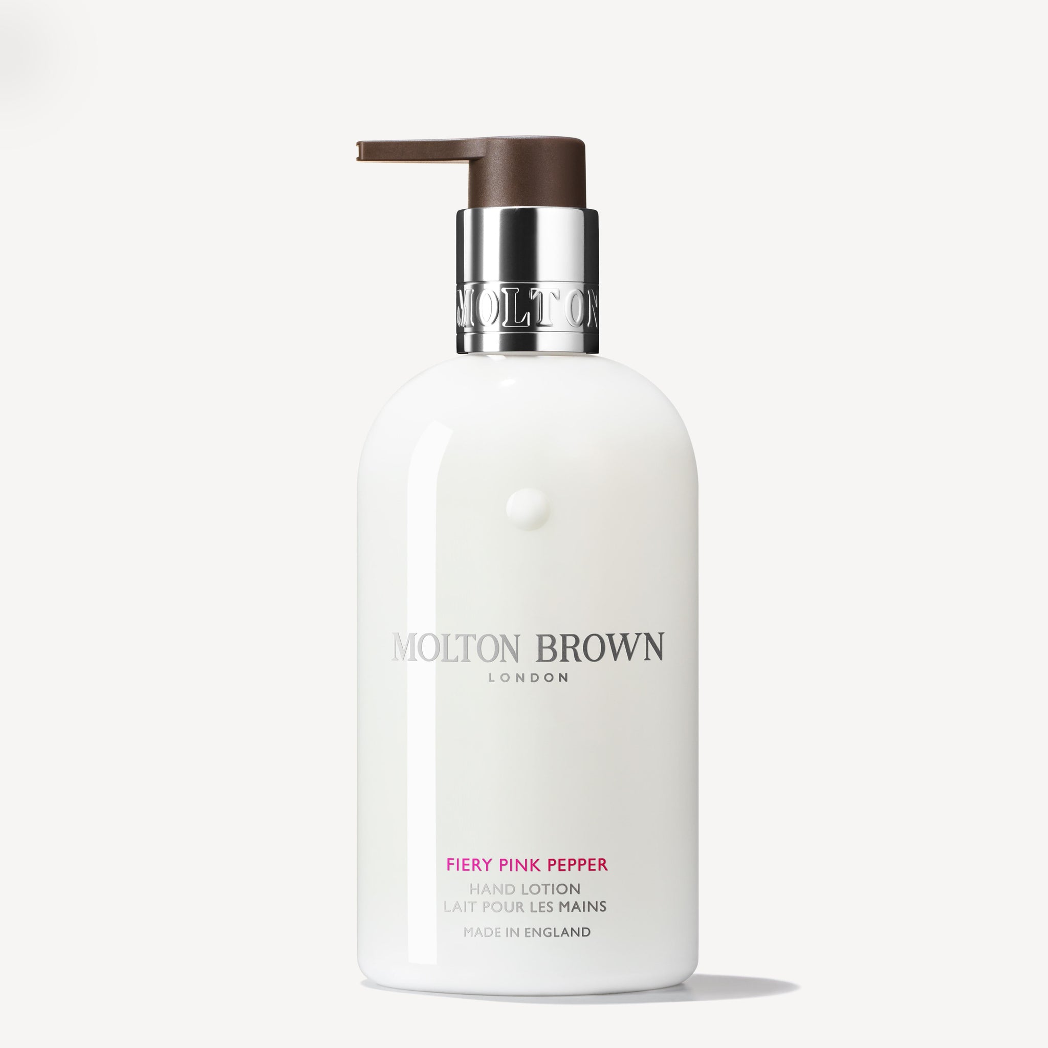 A white 300ml bottle of MoltonBrown Nederlands Fiery Pink Pepper Handlotion with a silver collar and brown pump, featuring silver and pink text. Provides hydrating care with subtle houtige noten.
