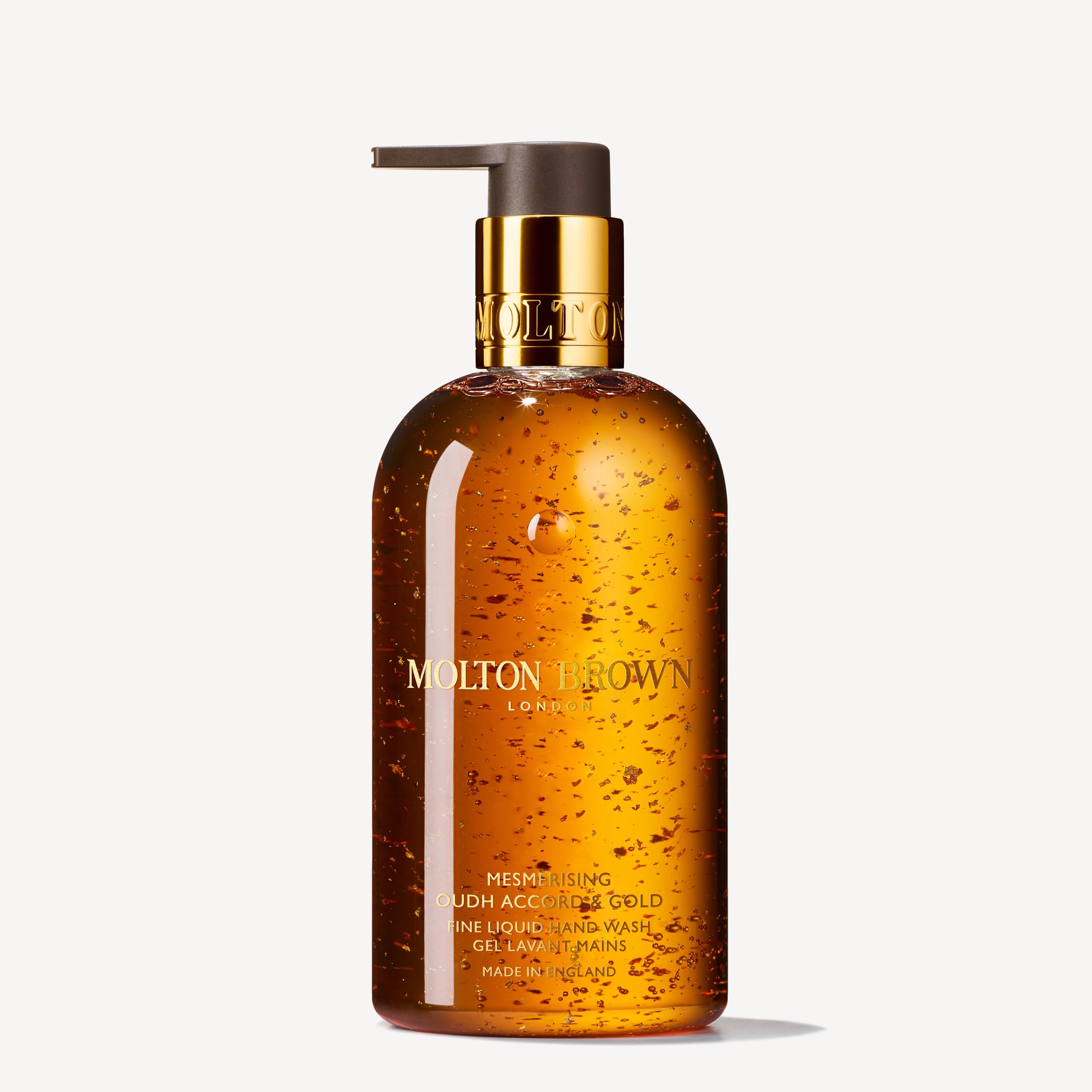 A clear pump bottle of MoltonBrown Nederlands Mesmerising Oudh Accord & Gold Vloeibare Handzeep 300ml, with luxe geur, amberachtige zeep en zichtbare gouddeeltjes. De elegante gouden pomp voegt een verfijnde touch toe.