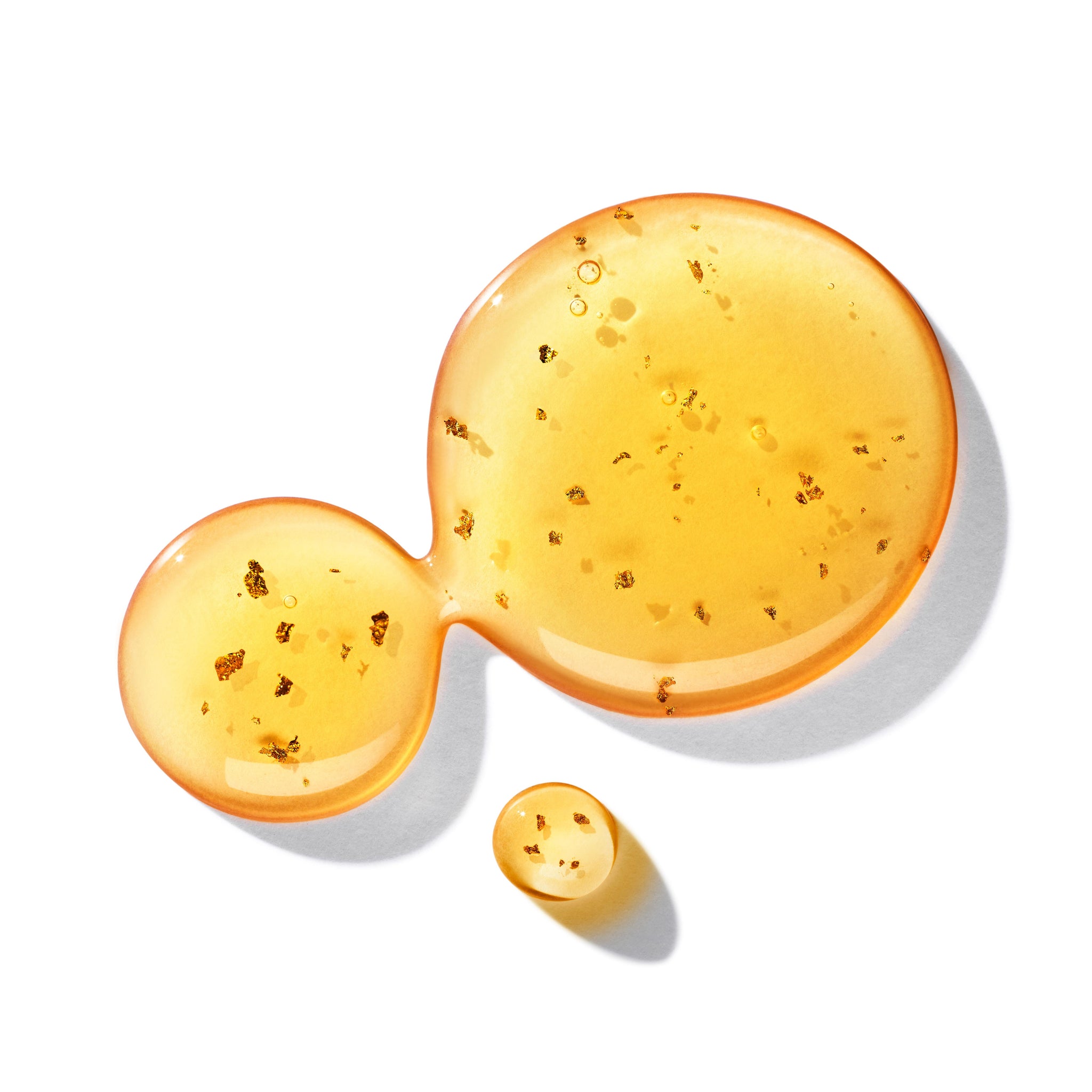 Three droplets of MoltonBrown Nederlands Mesmerising Oudh Accord & Gold Vloeibare Handzeep 300ml, flecked with gold, rest on white: two larger drops merge gracefully while a smaller one sits nearby, each casting soft shadows.