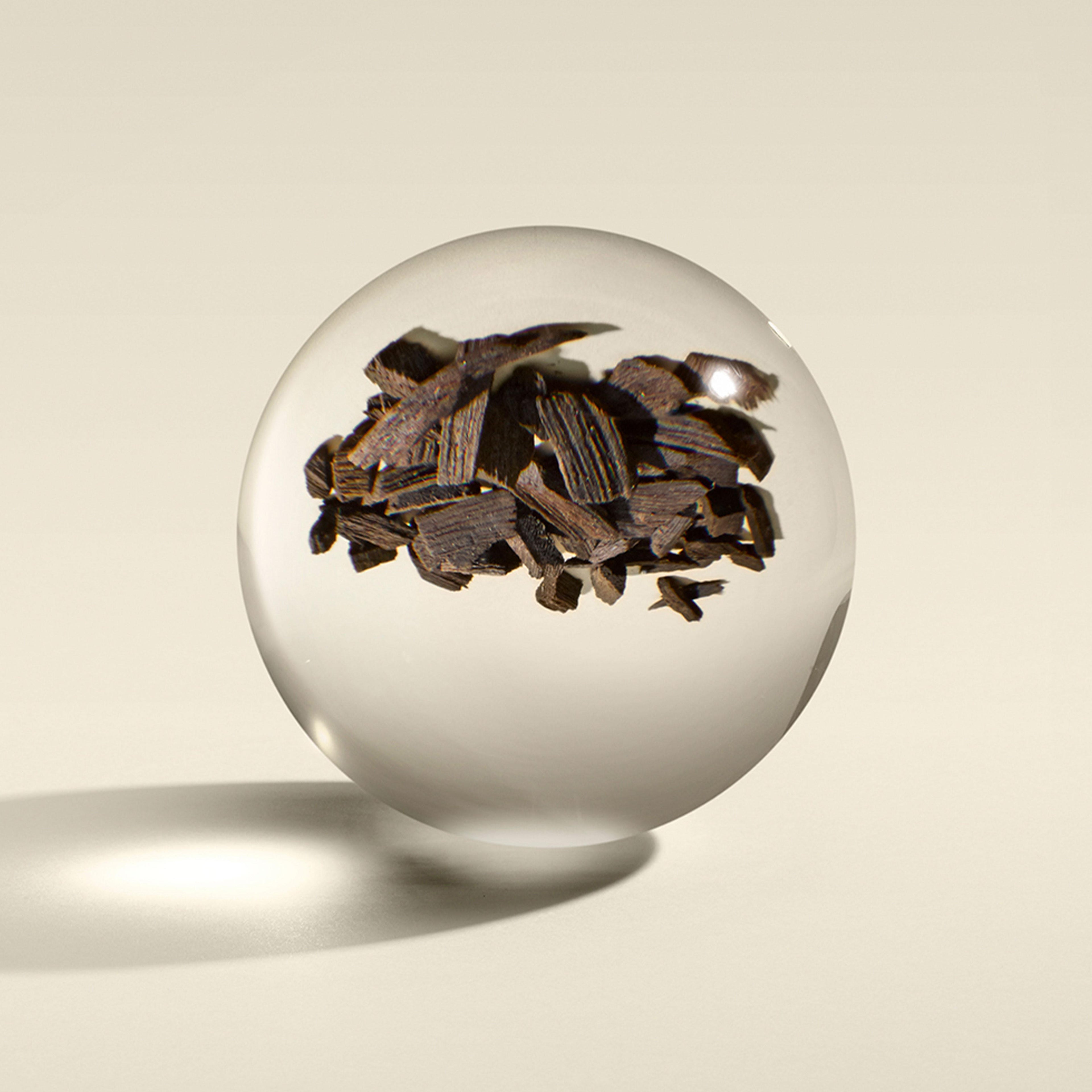 A clear glass sphere, set against a beige background, showcases the rich essence of Mesmerising Oudh Accord & Gold Vloeibare Handzeep 300ml by MoltonBrown Nederlands, capturing the luxury of deep oudh wood chips.