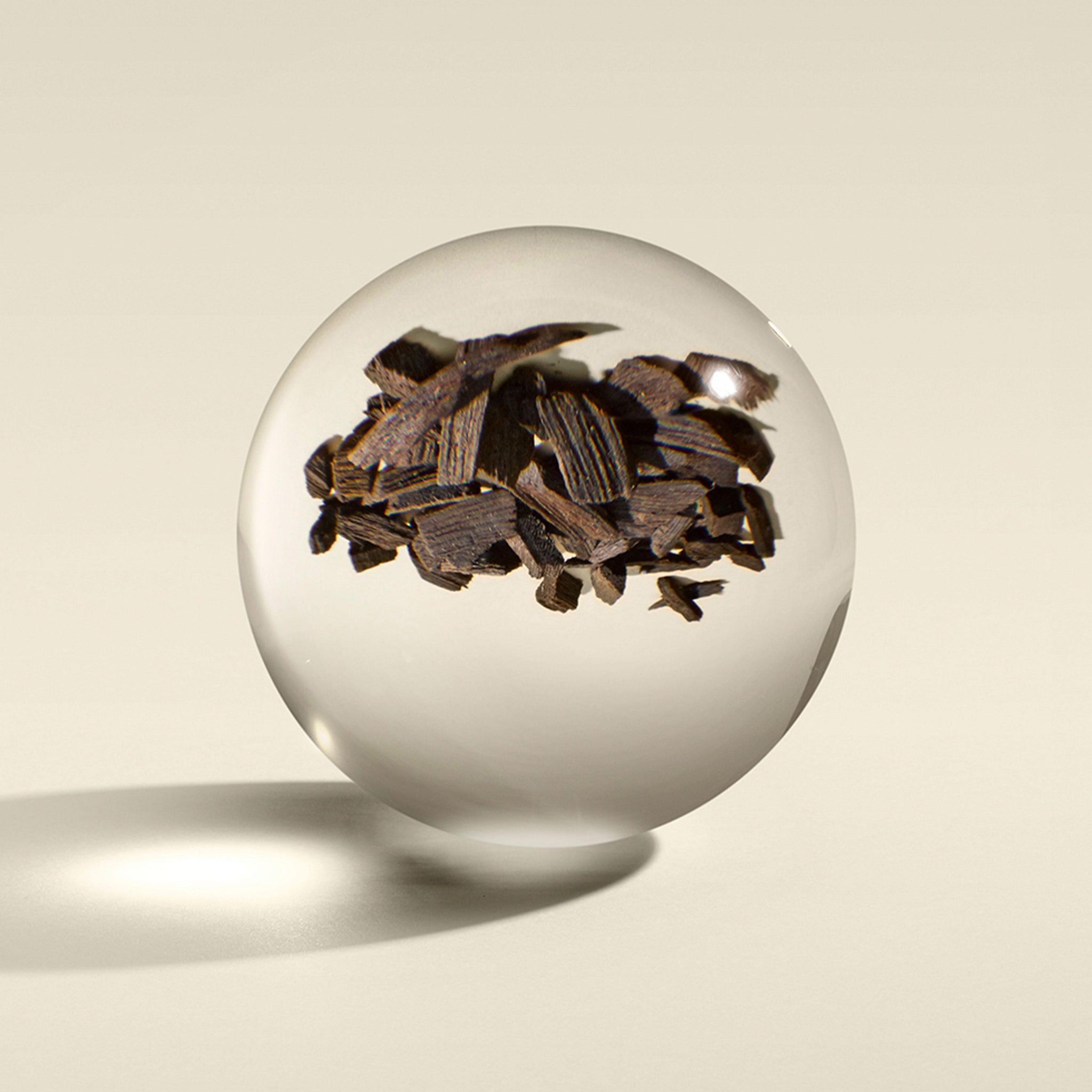 The Mesmerising Oudh Accord & Gold Handlotion 300ml by MoltonBrown Nederlands features a clear glass sphere with dark brown wood chips, evoking echte oudh, and casting a soft shadow on a light, neutral surface.