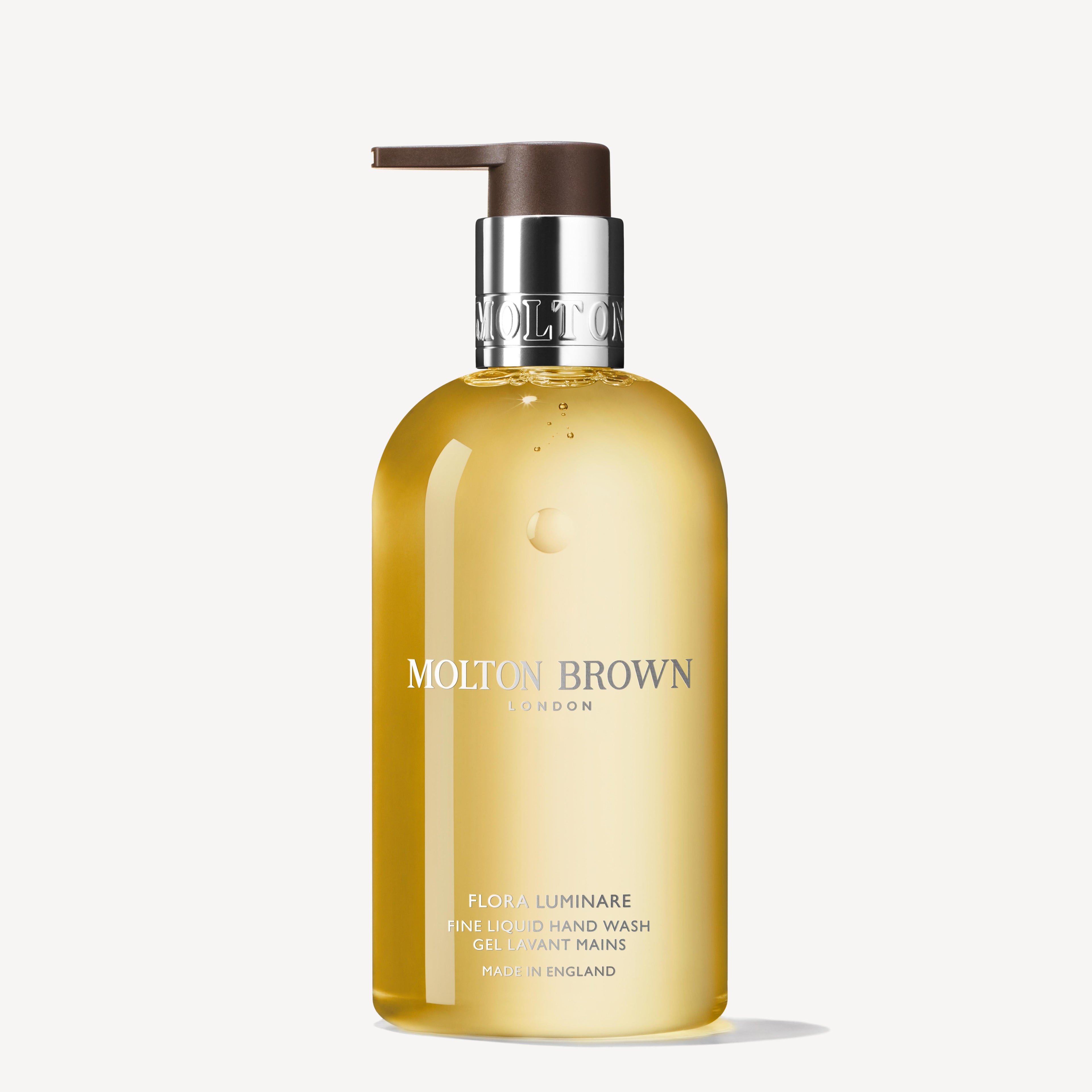 A clear bottle of MoltonBrown Nederlands Flora Luminare Vloeibare Handzeep 300ml with golden-yellow liquid, notes of sinaasappelbloesem and cederhout, silver collar, dark pump dispenser, upright on a white background.