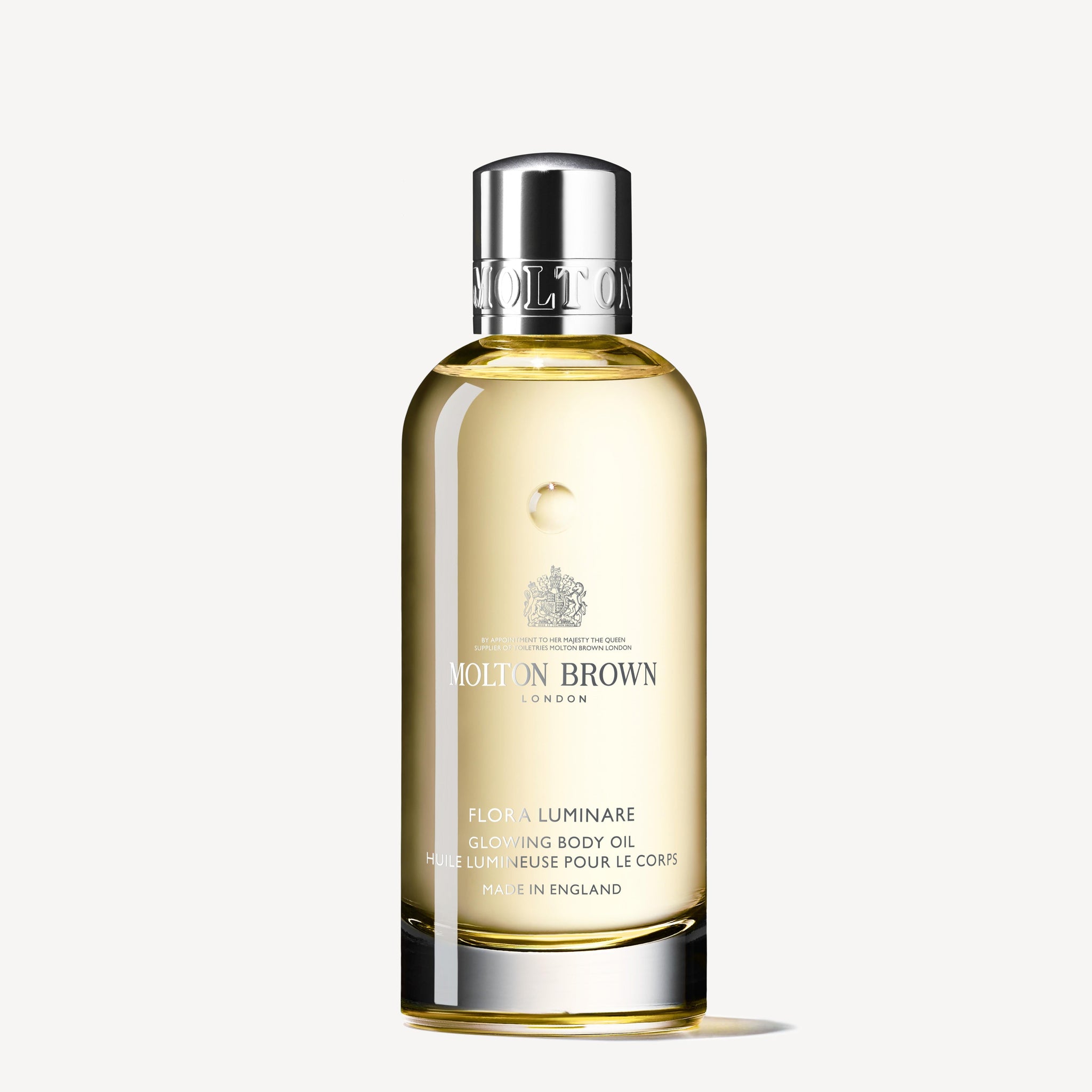 A clear bottle of MoltonBrown Nederlands Flora Luminare Glowing Lichaamsolie 100ml with a silver cap, filled with light golden oil, is shown against a plain white background.