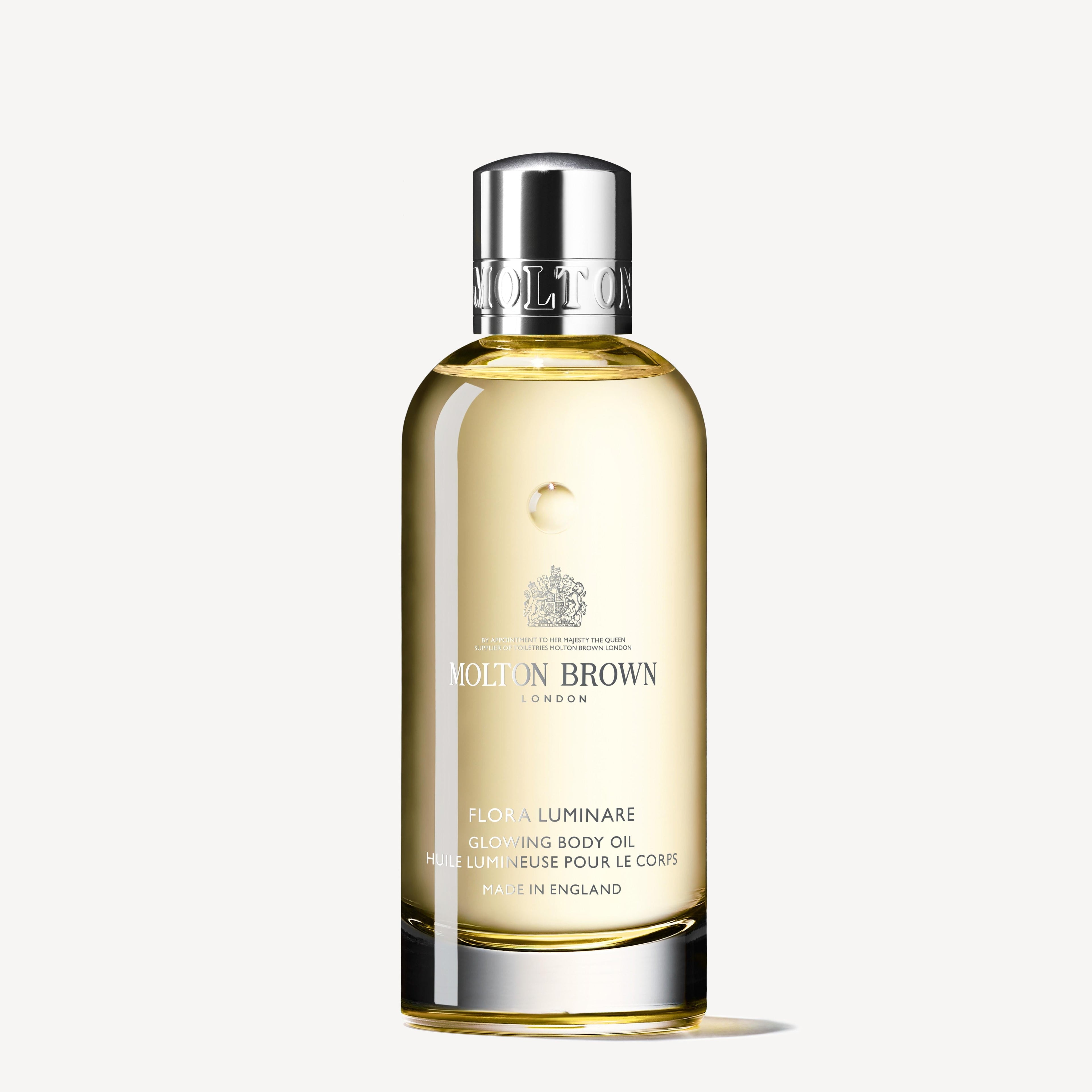A clear bottle of MoltonBrown Nederlands Flora Luminare Glowing Lichaamsolie 100ml with a silver cap, filled with light golden oil, is shown against a plain white background.
