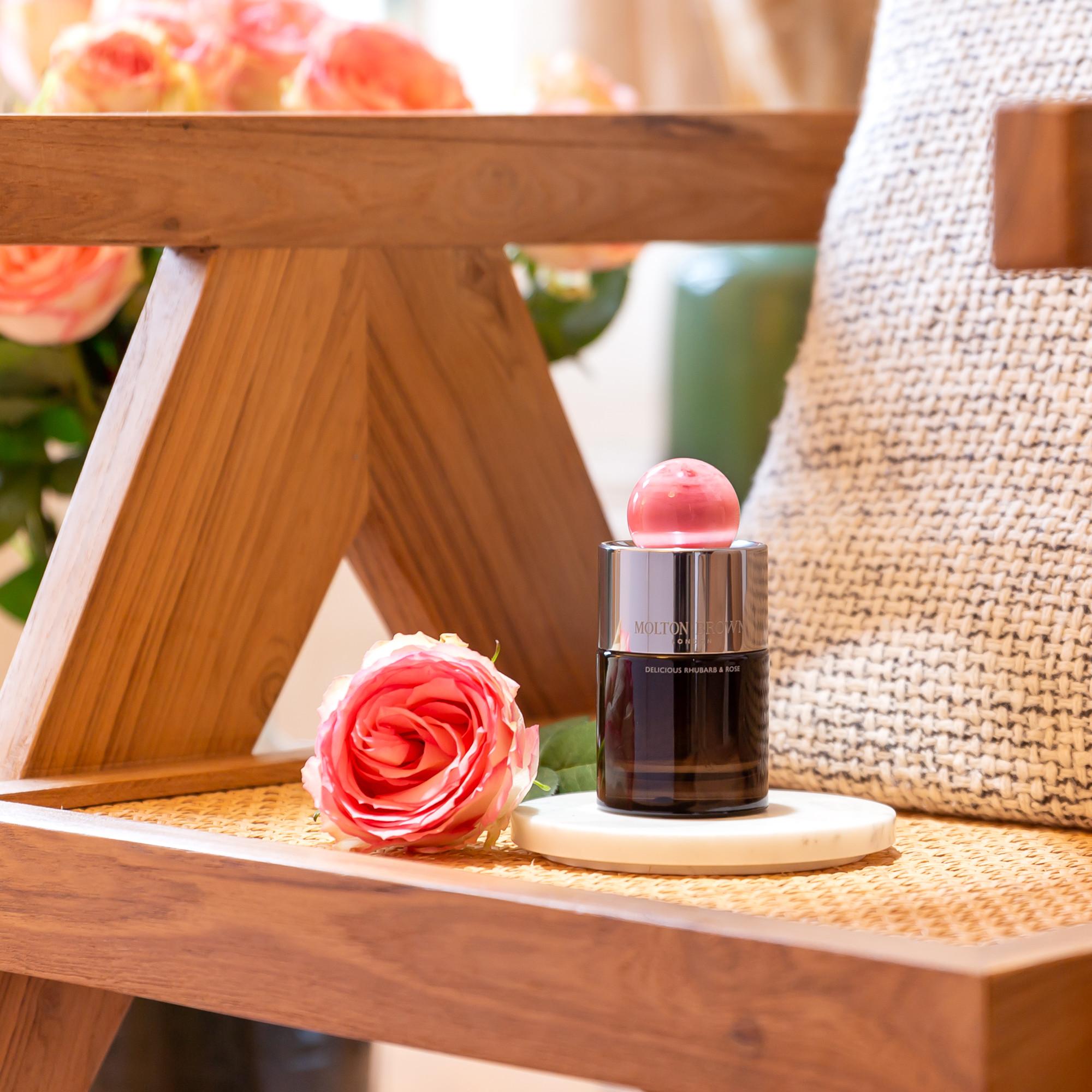 A bottle of MoltonBrown Nederlands Delicious Rhubarb & Rose Eau de Parfum 100ml with a pink cap sits beside a pink rose on a wooden chair, evoking the gentle scent of jasmijn and the elegance of fresh roses in bloom.