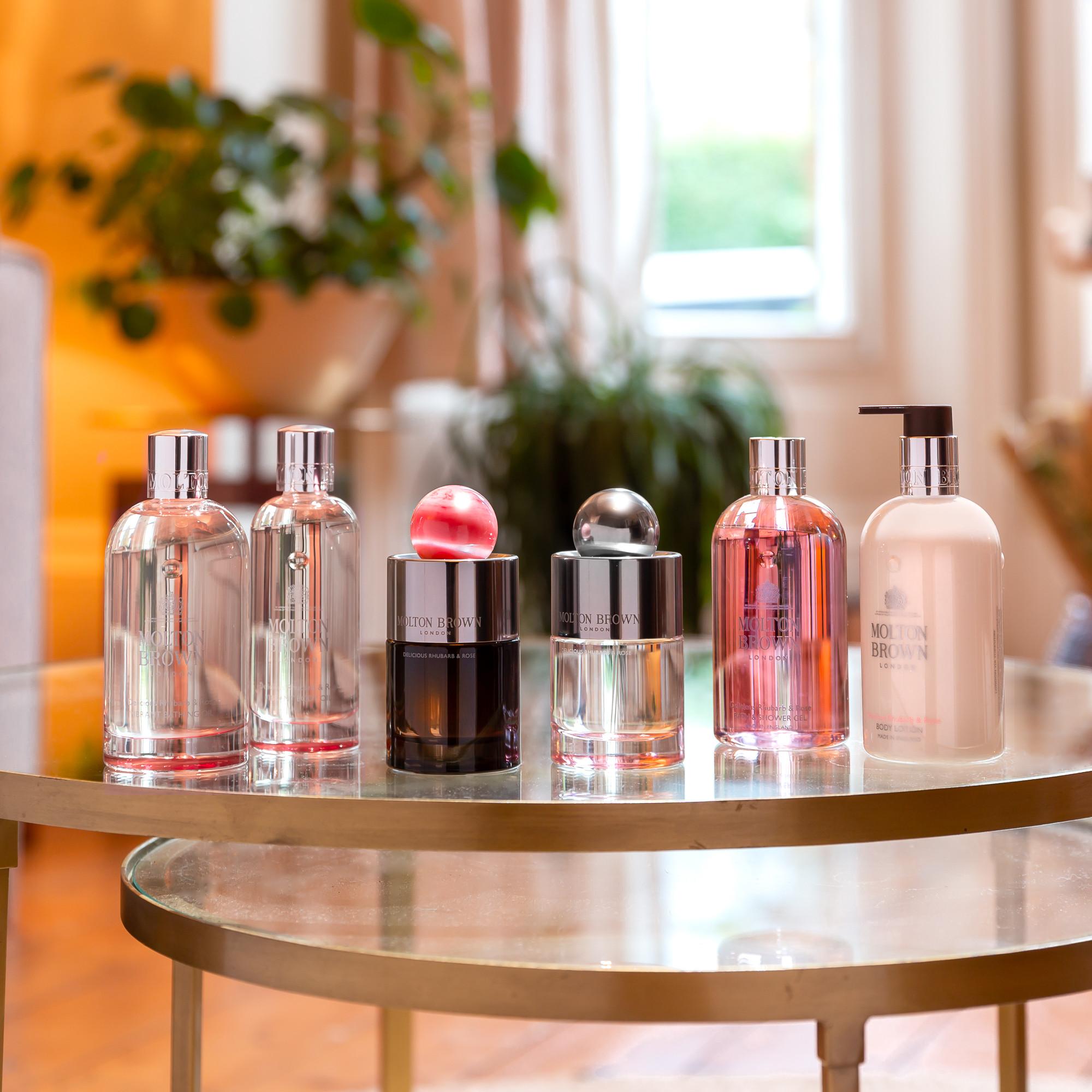 Five MoltonBrown Nederlands Delicious Rhubarb & Rose Eau de Parfum 100ml bottles are arranged on a glass table in a sunlit, plant-filled living room, filling the space with elegance and a delicate fragrance.