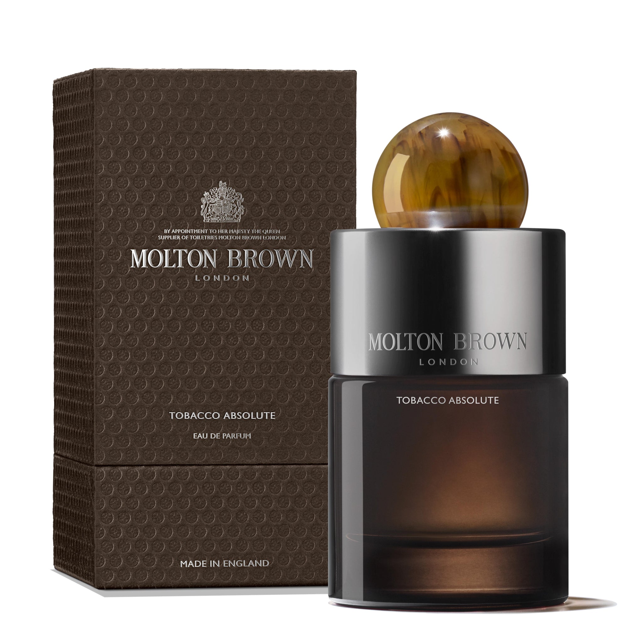 A 100ml bottle of MoltonBrown Nederlands Tobacco Absolute Eau de Parfum stands by its textured brown box, showcasing a round marbled cap, silver accents, and notes of cederhout and sinaasappelbloesem.
