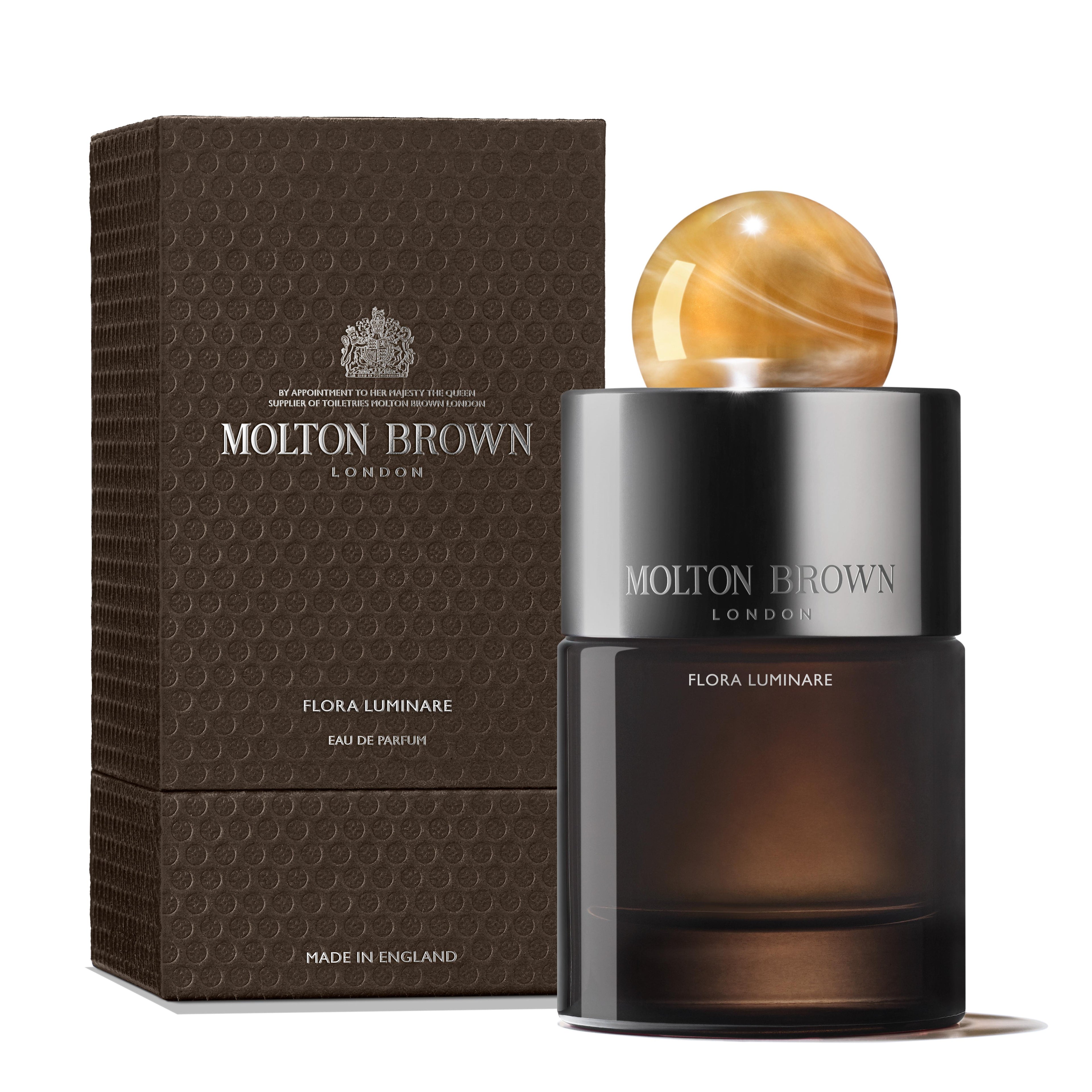 A 100ml Flora Luminare Eau de Parfum by MoltonBrown Nederlands, featuring a round golden marble-effect cap, is shown beside its textured dark brown box. Both bottle and packaging display the product name and branding.