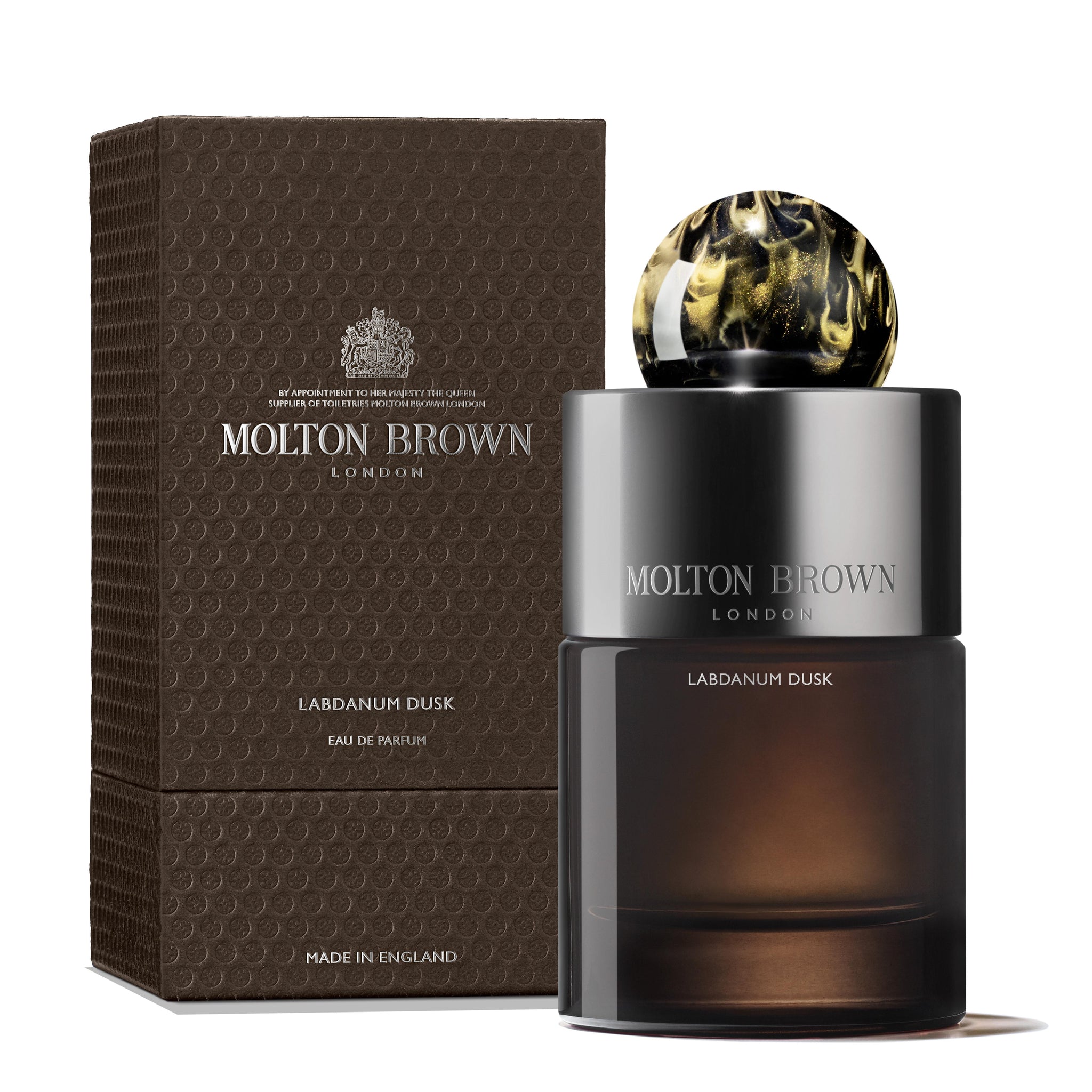 A bottle of MoltonBrown Nederlands Labdanum Dusk Eau de Parfum 100ml stands beside its textured brown box. The dark, translucent bottle has cedarwood notes, a shiny silver label, and a round marbled black-and-gold cap.
