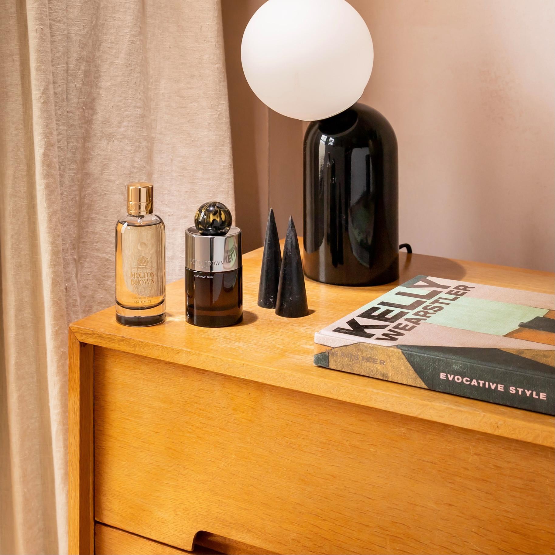 A wooden dresser holds two MoltonBrown Nederlands Labdanum Dusk Eau de Parfum 100ml bottles, two black cone-shaped objects, a black lamp with a round white shade, and a book titled KELLY WEARSTLER. Beige curtains hang in the background.