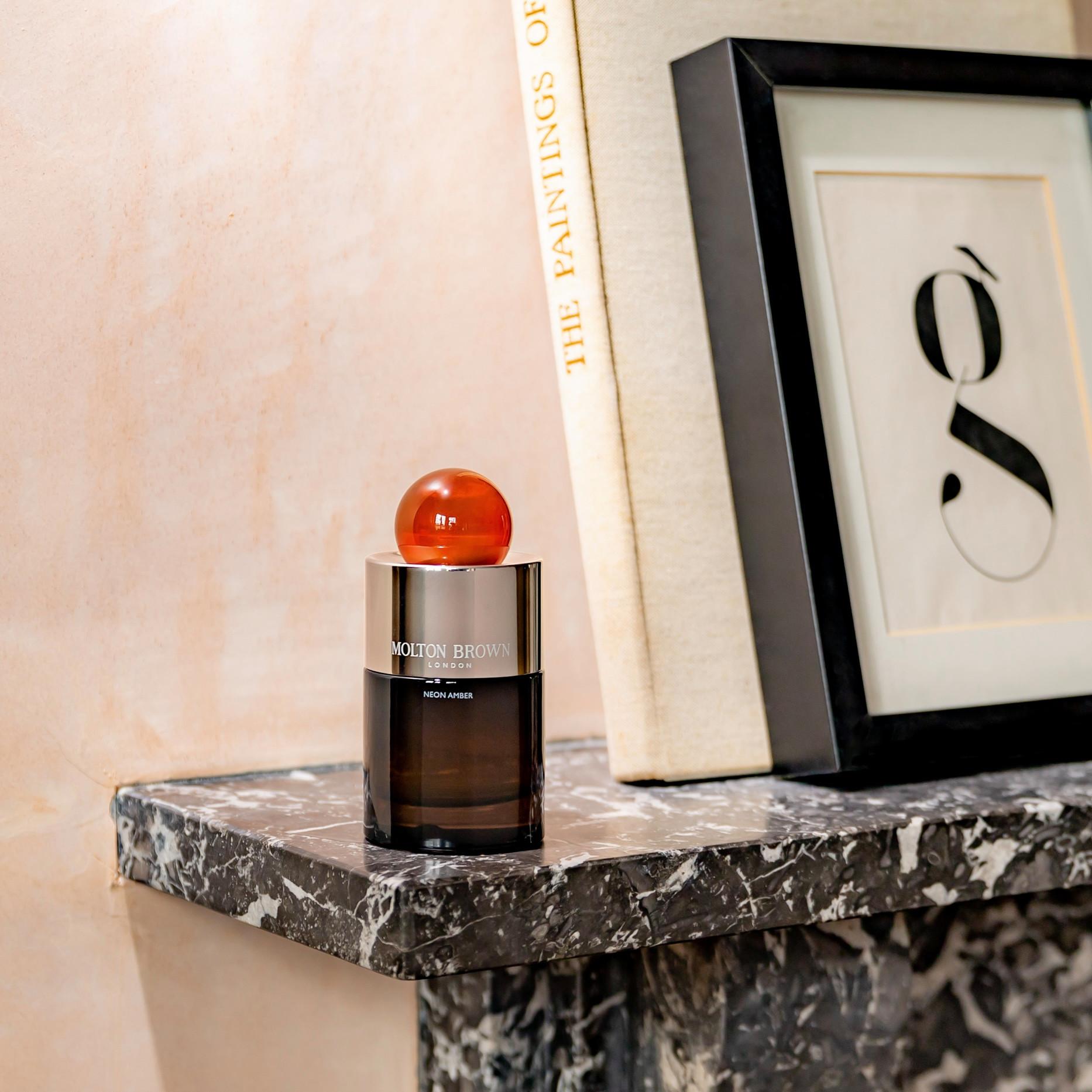 A bottle of MoltonBrown Nederlands Neon Amber Eau de Parfum 100ml with a round orange cap sits on a black marble shelf beside a framed artwork featuring a stylized letter g and the book The Paintings of.