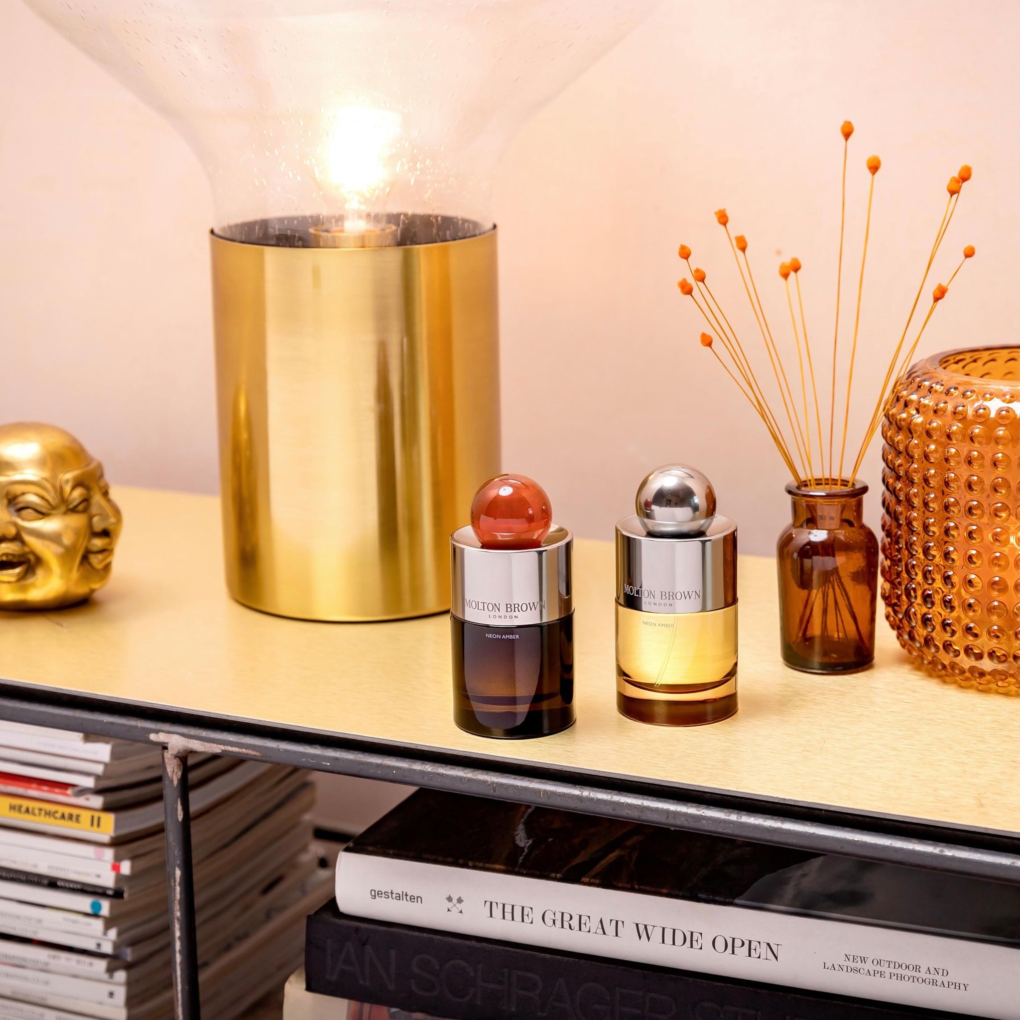 A modern tabletop styled with two MoltonBrown Nederlands Neon Amber Eau de Parfum 100ml bottles, a gold lamp, small vase of orange stems, amber jar, and gold face decor. Warm lighting and books on the lower shelf create a stylish scene.