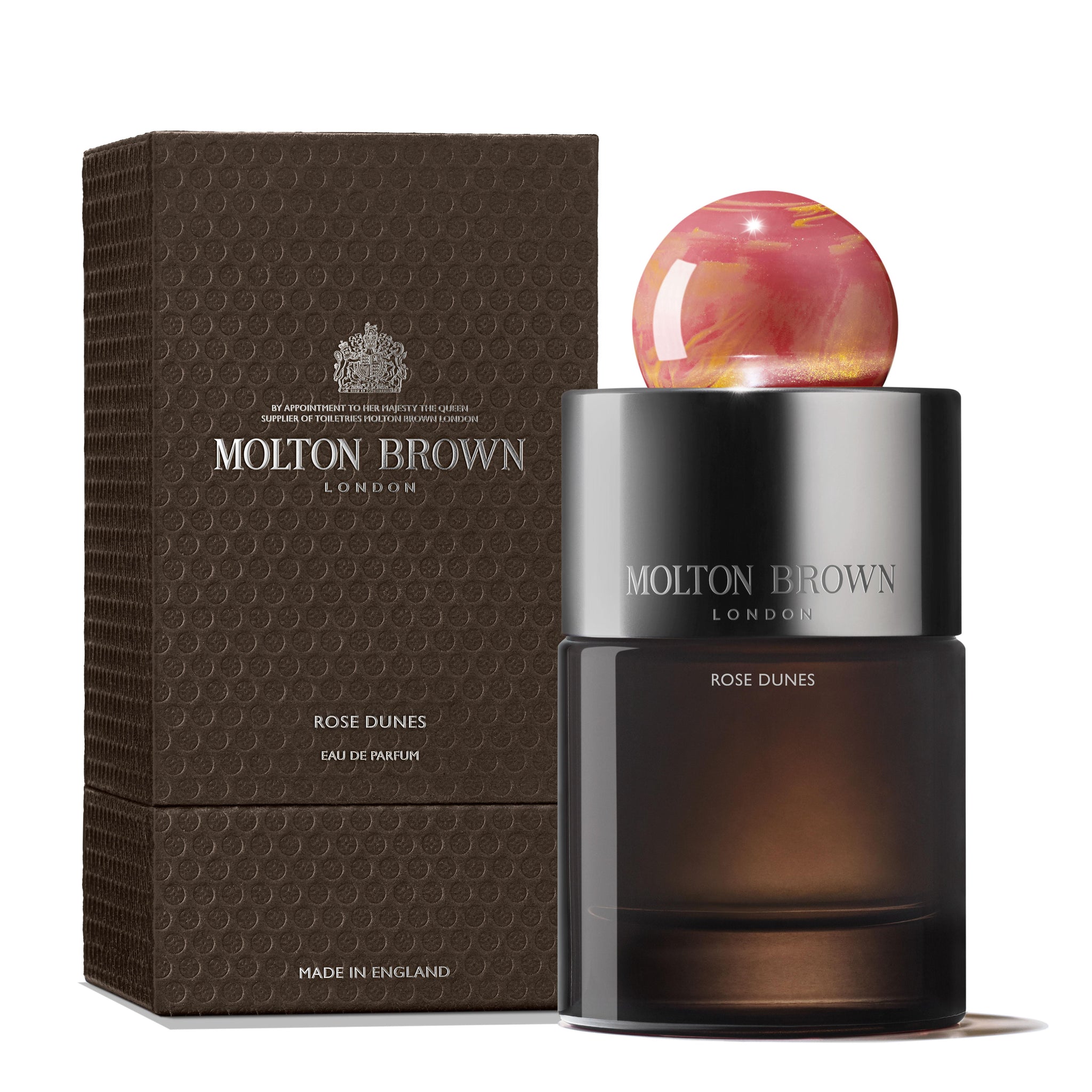 A bottle of MoltonBrown Nederlands Rose Dunes Eau de Parfum 100ml sits by its textured brown box, featuring a gradient amber base and marble-effect cap that mirror the fragrance’s rich amber patchouli scent and elegant style.