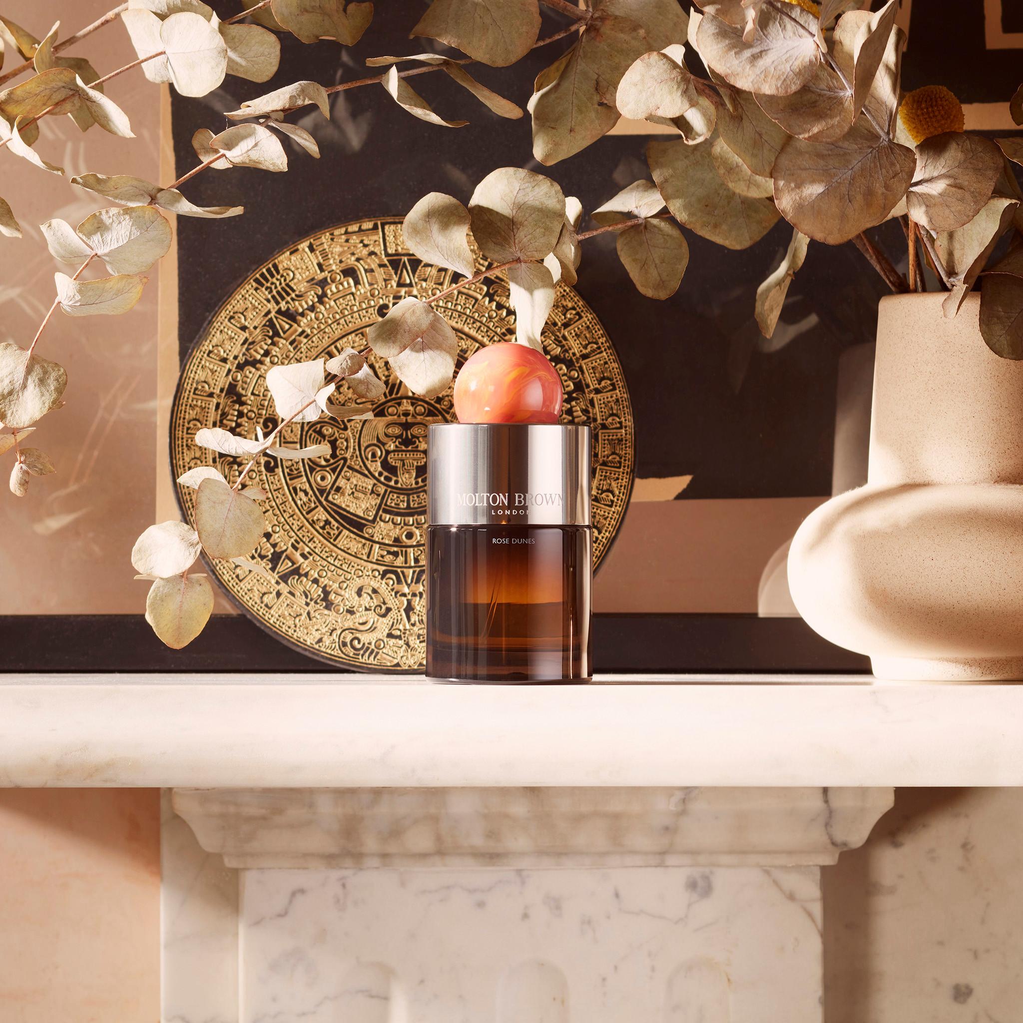 A brown glass bottle of MoltonBrown Nederlands Rose Dunes Eau de Parfum 100ml rests on a marble shelf, surrounded by dried eucalyptus branches, a gold-patterned round plate, and a beige ceramic vase.