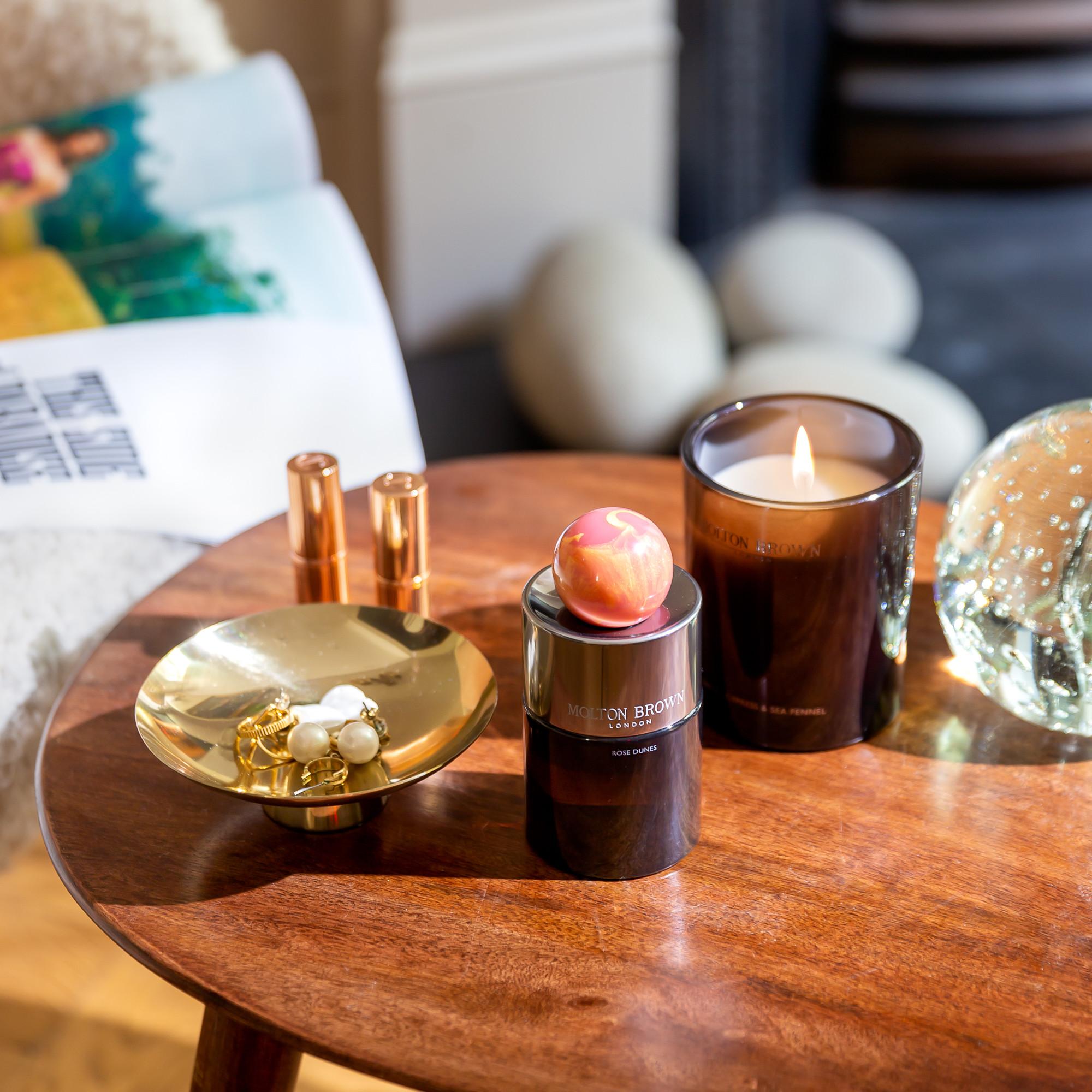 A wooden table holds a lit candle, a MoltonBrown Nederlands Rose Dunes Eau de Parfum 100ml bottle with a decorative object, a gold dish with jewelry, two lipsticks, and a glass ornament. A blurred magazine and soft furnishings are in the background.