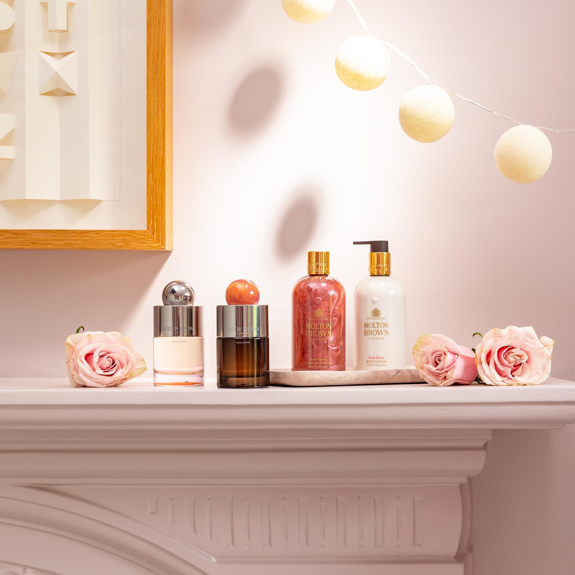 A mantel topped with pink roses, a tray holding two MoltonBrown Nederlands bottles—including Rose Dunes Eau de Parfum 100ml—plus two more perfumes, all against a soft pink wall with string lights and framed art.