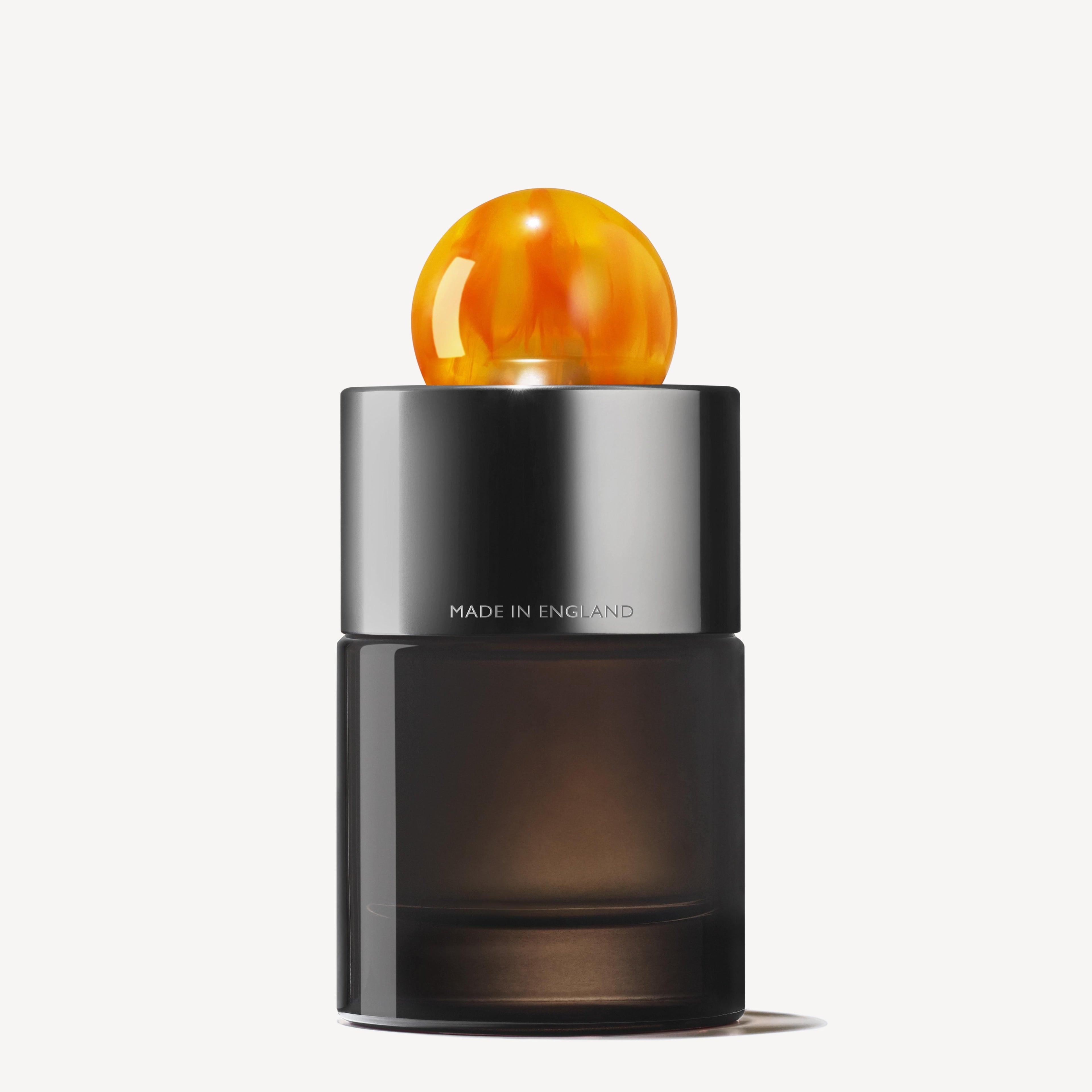 A sleek, dark glass bottle of MoltonBrown Nederlands Sunlit Clementine & Vetiver Eau de Parfum 100ml features a shiny silver cap with a round amber topper. "MADE IN ENGLAND" is visible, set against a plain white background.