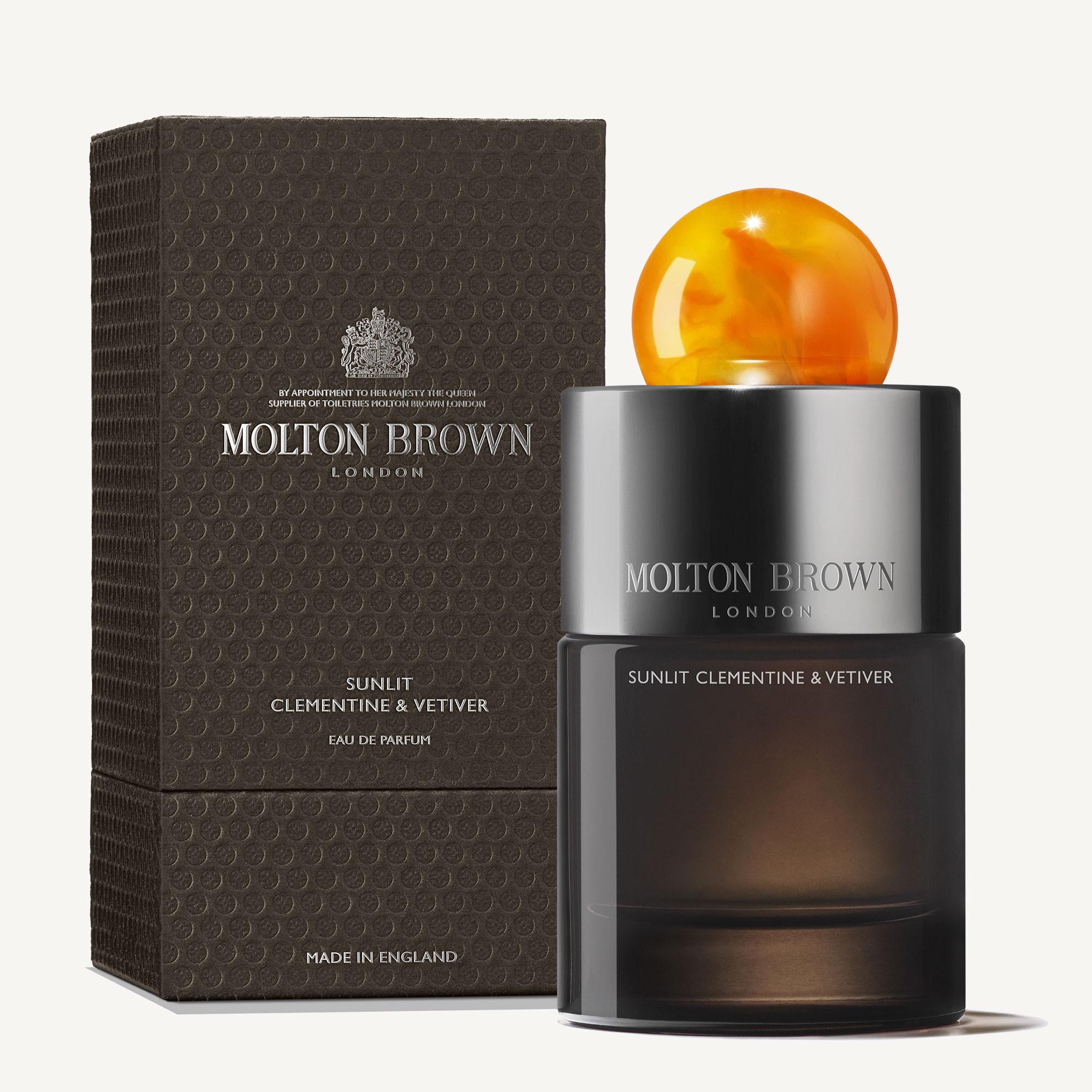 A 100ml bottle of MoltonBrown Nederlands Sunlit Clementine & Vetiver Eau de Parfum with a round amber cap stands by its textured dark box, both showcasing the brand and inviting you to discover its long-lasting patchouli notes.