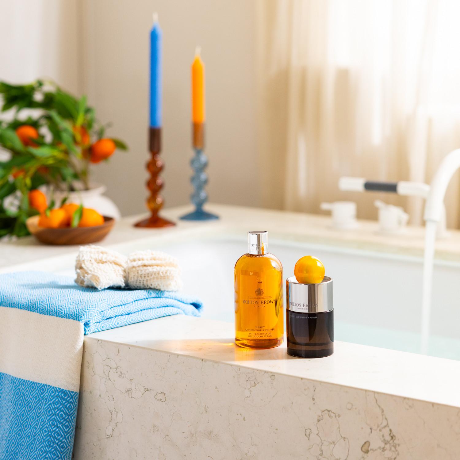 The MoltonBrown Nederlands Sunlit Clementine & Vetiver Eau de Parfum 100ml sits by a marble tub, surrounded by blue towels, candles, and fresh oranges for a patchouli-infused, spa-like vibe.