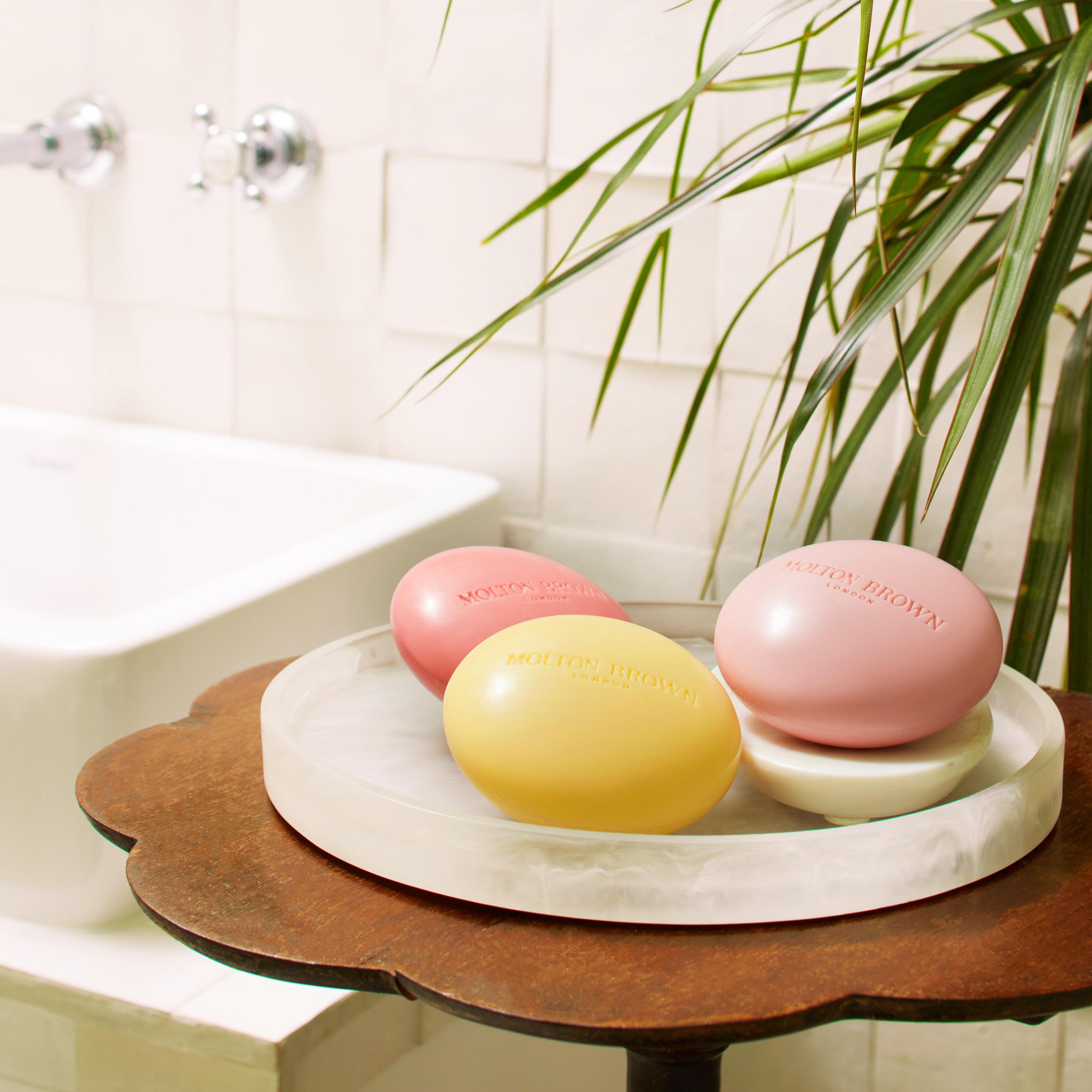 Three pastel Orange & Bergamot Perfumed Soap bars by MoltonBrown Nederlands, each 150g and enriched with natuurlijke oliën, rest on a white tray atop wood in a bathroom with tiled walls, a white tub, and nearby green leaves.