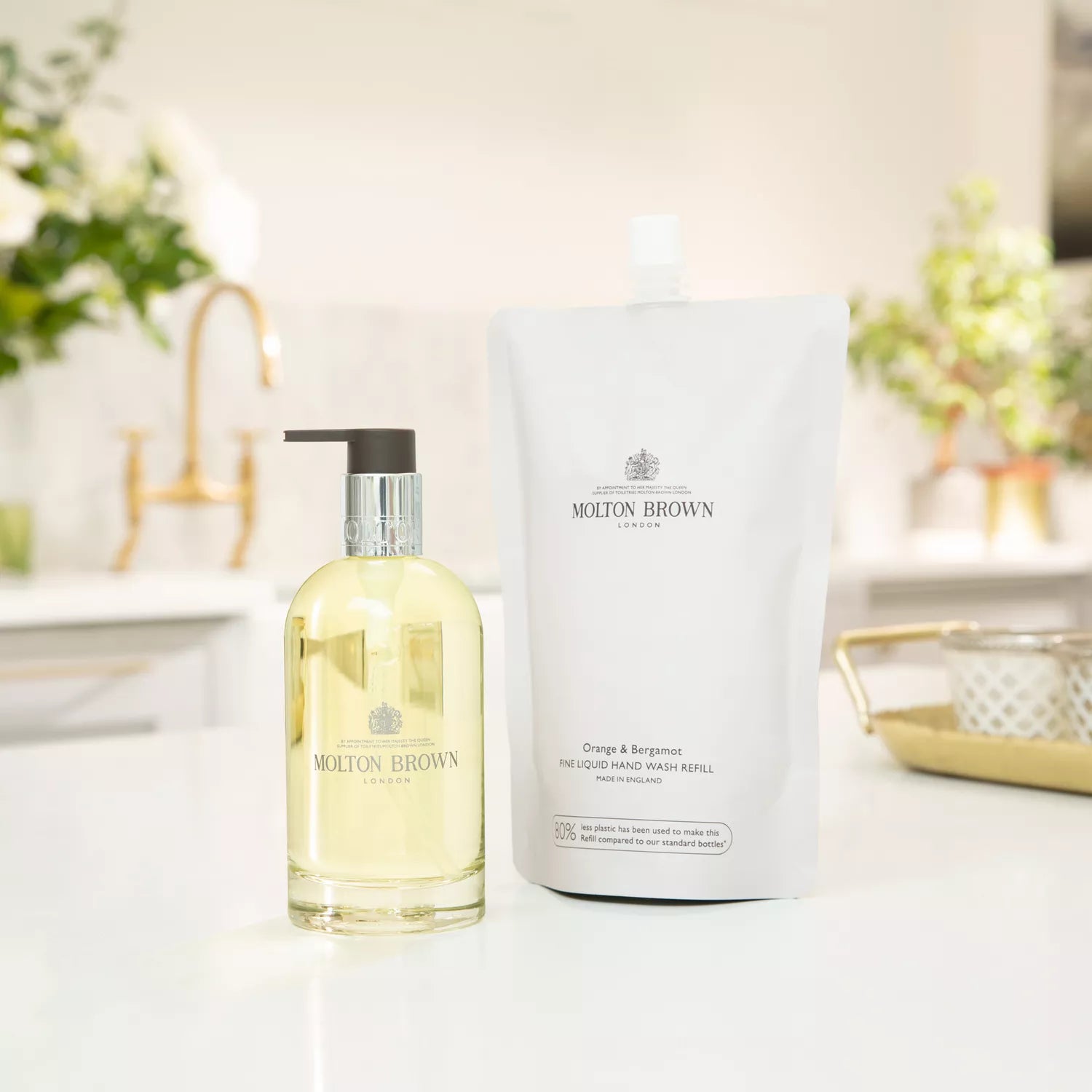 Een MoltonBrown Nederlands Orange & Bergamot Vloeibare Handzeep Glazen Fles 200ml met pomp staat naast een refill pouch op een licht keukenblad, met vage planten en keukenitems op de achtergrond.
