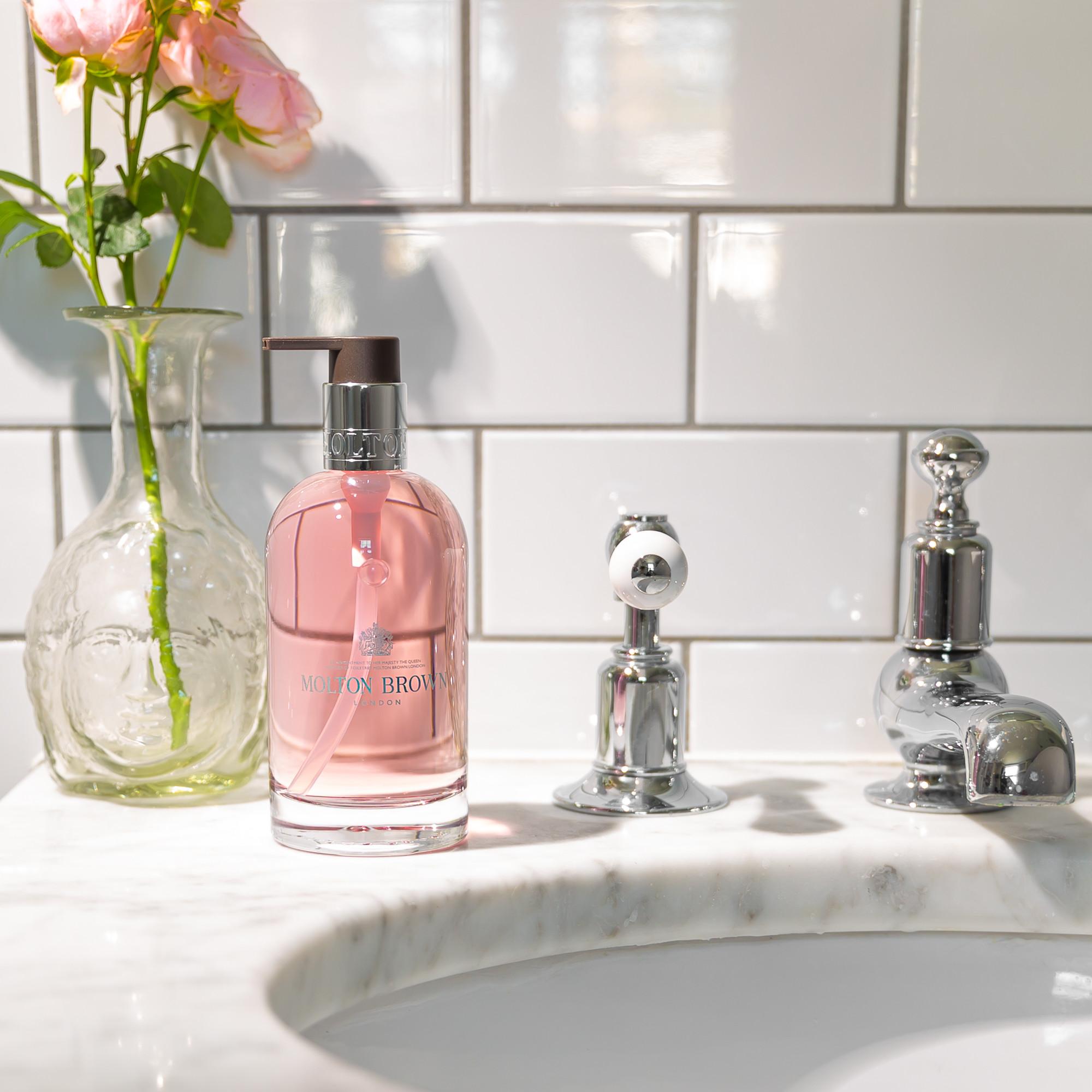 A 200ml MoltonBrown Nederlands Delicious Rhubarb & Rose Vloeibare Handzeep Glazen Fles sits on a marble sink beside a chrome faucet, with luxe verzorgingsproducten and pink flowers against a white tiled wall.