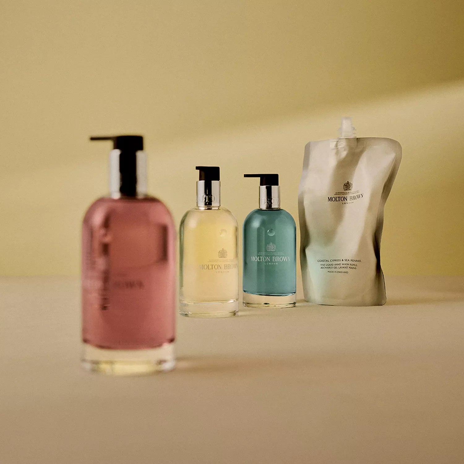 Four MoltonBrown Nederlands Coastal Cypress & Sea Fennel Vloeibare Handzeep Glazen Fles 200ml bottles, filled with pink, yellow, and teal liquids, stand before a silver refill pouch on a beige background.