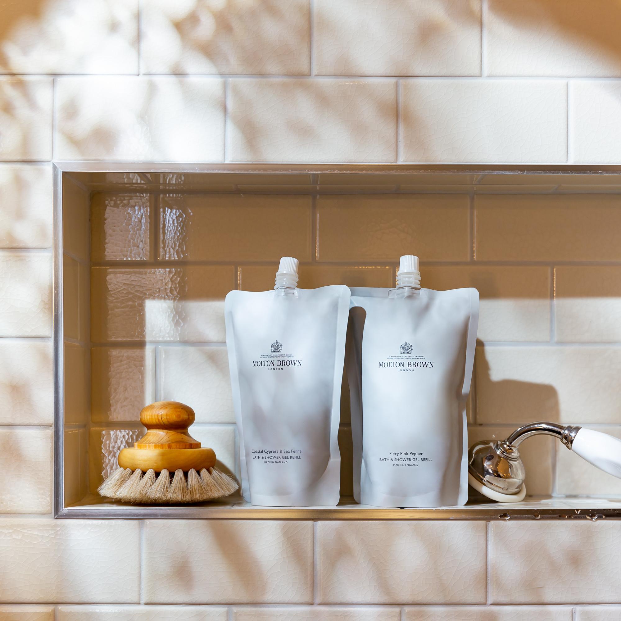 A tiled bathroom niche holds two Fiery Pink Pepper Bad- & Douchegel Navulling 400ml pouches from MoltonBrown Nederlands, a wooden-handled body brush, and a chrome showerhead. Soft light and shadows fall across the scene.