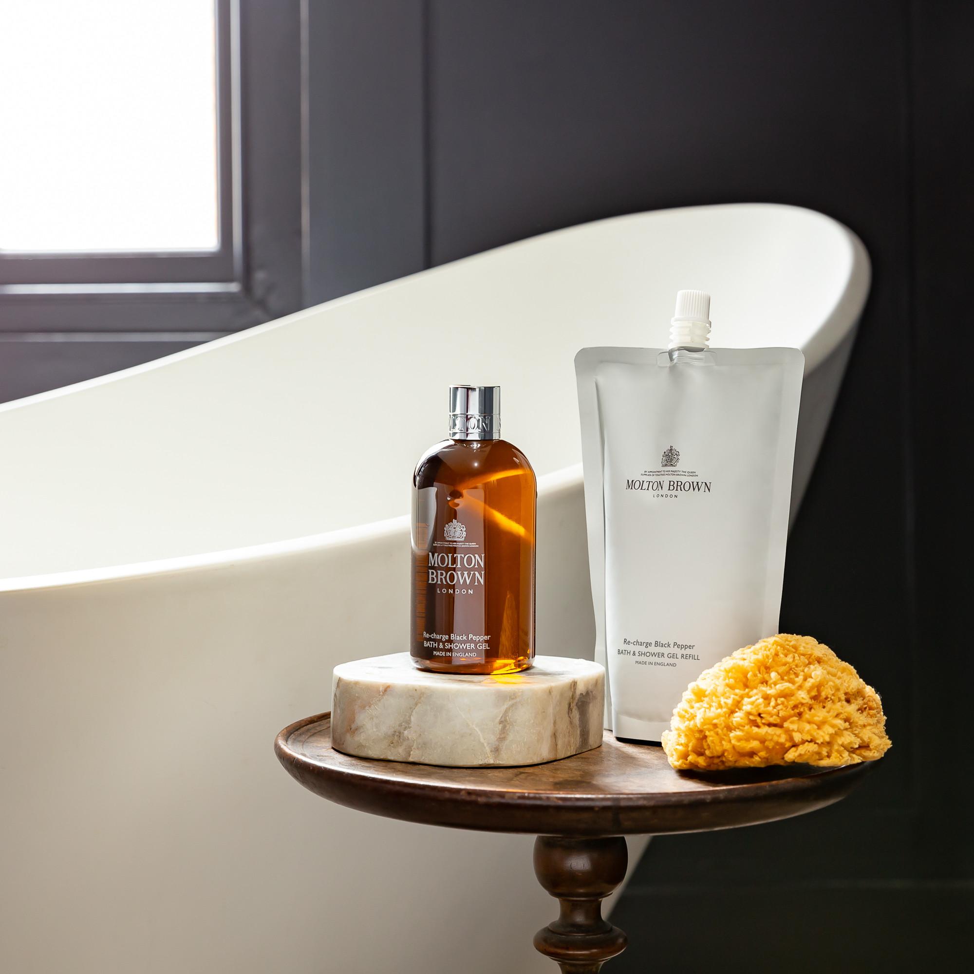 MoltonBrown Nederlands' Re-charge Black Pepper Bad- & Douchegel Navulling 400ml and a natural sponge are displayed on a small round table next to a white bathtub, with a dark wall and window in the background.