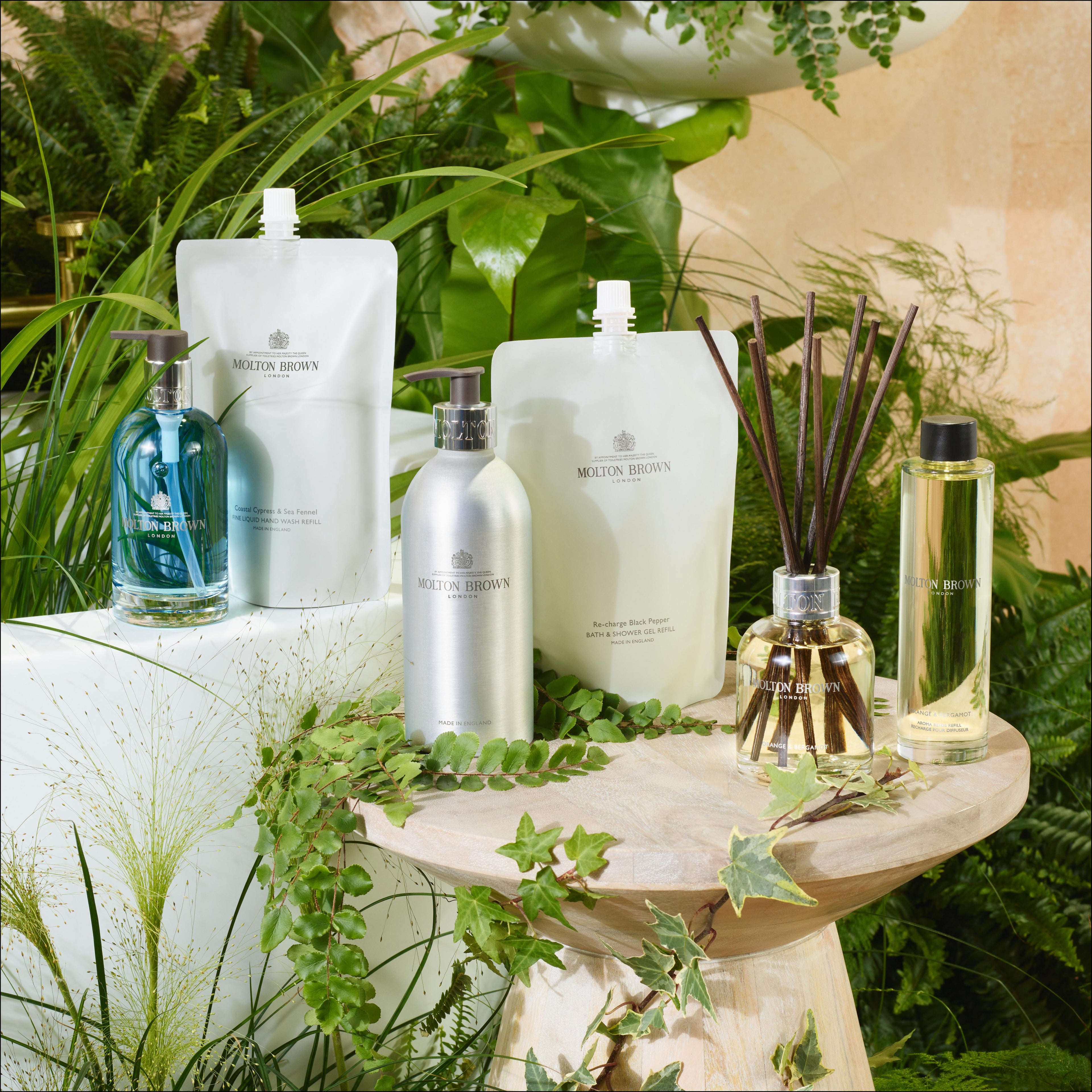 MoltonBrown Nederlands Coastal Cypress & Sea Fennel Navulbare Fles 400ml staat op een houten kruk, omringd door weelderige groene planten en bladeren.