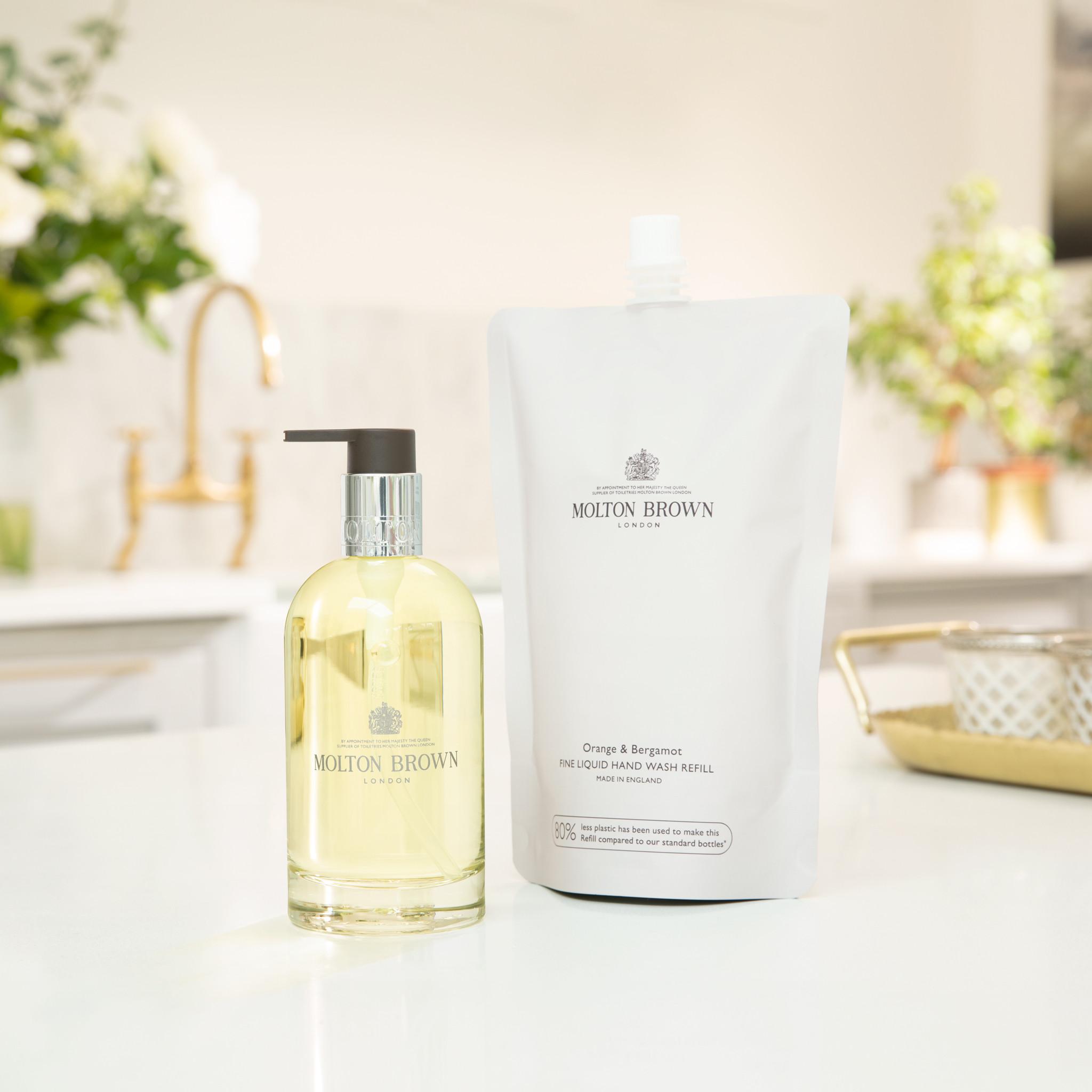 Een fles MoltonBrown Nederlands Orange & Bergamot Vloeibare Handzeep met pomp staat naast de 400ml navulling op een witte aanrecht in een lichte, moderne keuken, met een frisse citrusgeur en subtiele bloemen op de achtergrond.
