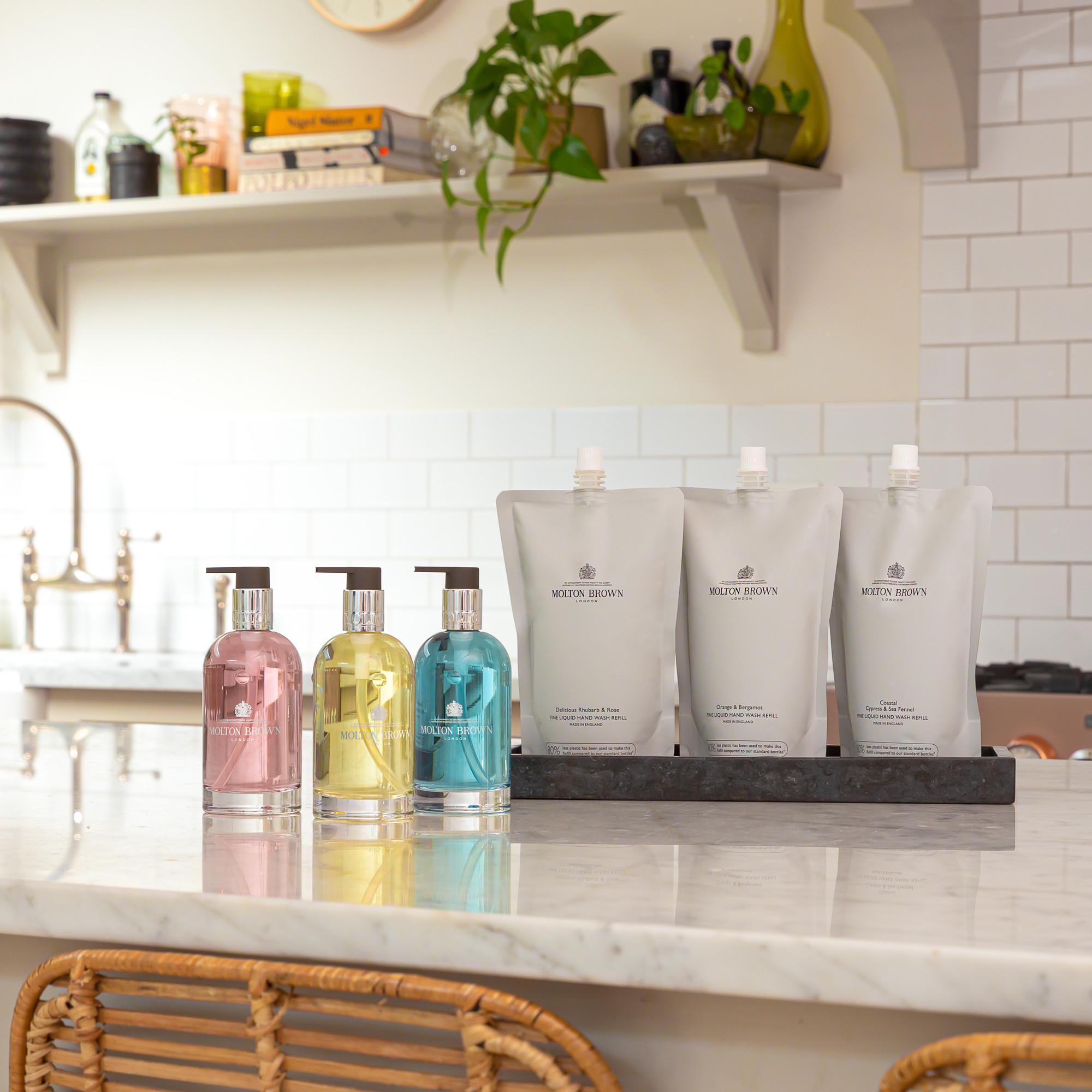 On a marble kitchen counter are three MoltonBrown Nederlands Orange & Bergamot Vloeibare Handzeep Navulling 400ml pouches with a fresh citrus scent, alongside colored dispensers; shelves behind hold books, plants, and kitchen items against tiled walls.