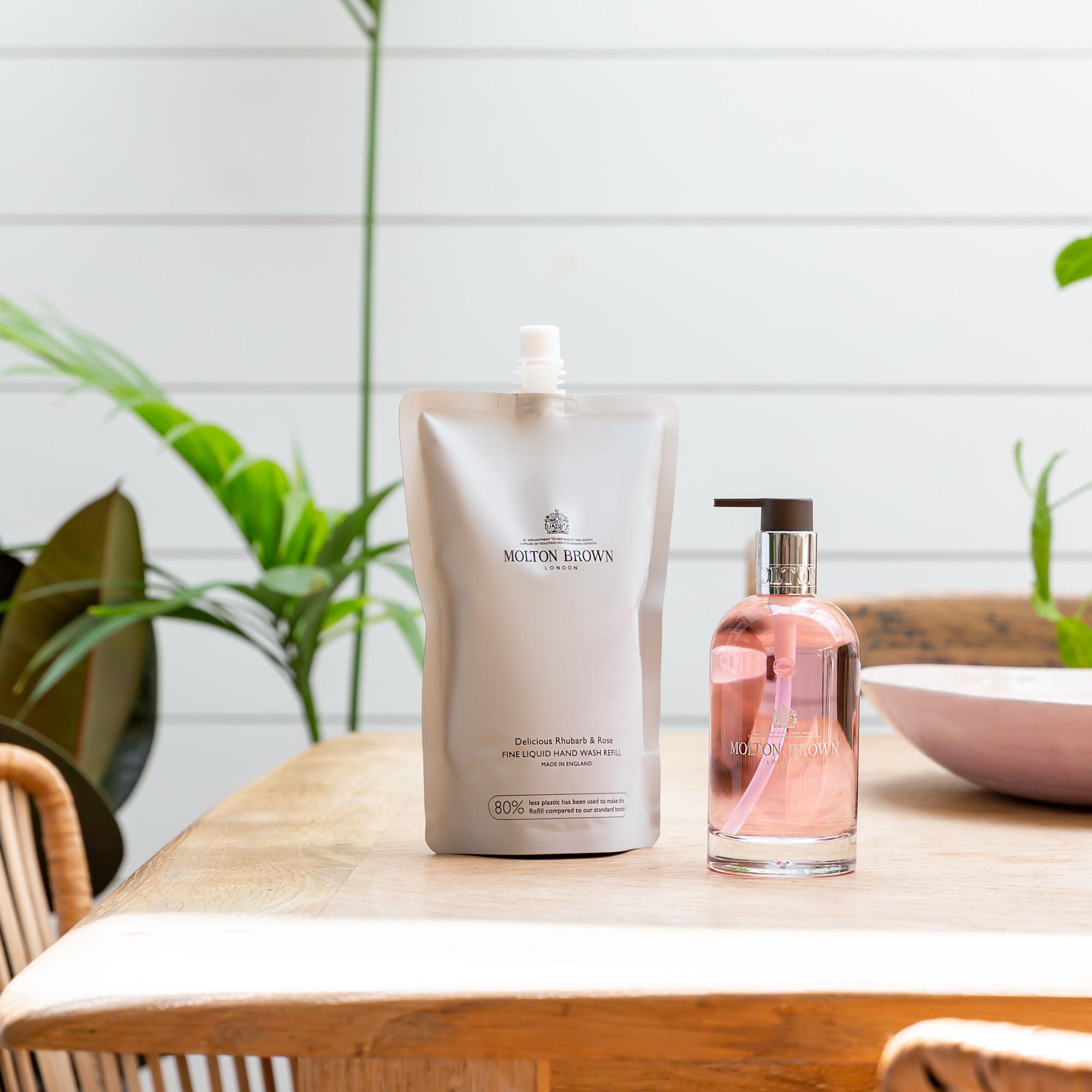 A MoltonBrown Nederlands Delicious Rhubarb & Rose Vloeibare Handzeep Navulling 400ml and a clear pump bottle with pink liquid rest on a light wooden table, surrounded by green plants. The bloemig-fruitige geur fills the bright, modern room.