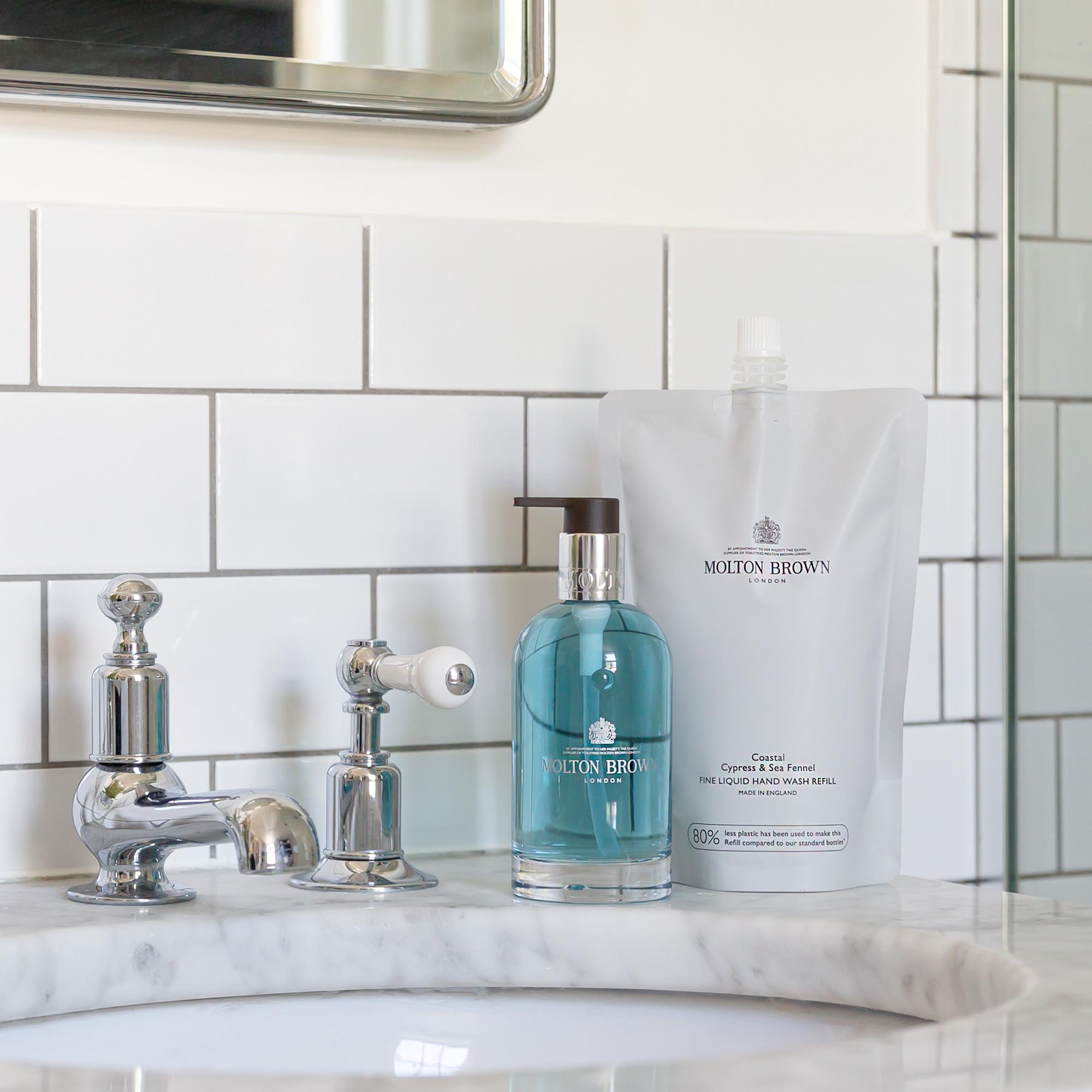 A bottle of MoltonBrown Nederlands soap and a Coastal Cypress & Sea Fennel Vloeibare Handzeep Navulling 400ml pouch rest on a marble sink by a chrome faucet—een milieuvriendelijke keuze met een aromatische geur.