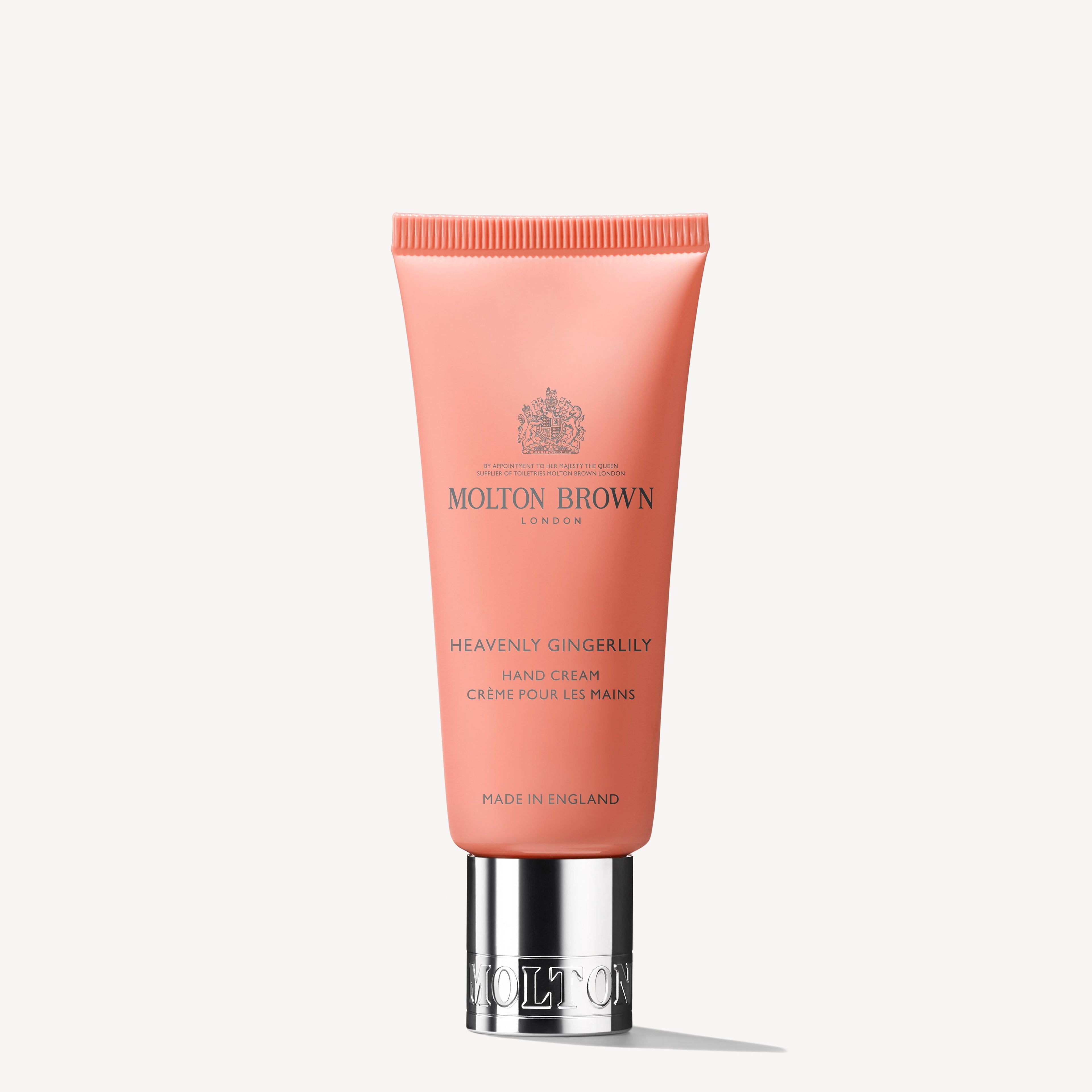 A coral pink tube of MoltonBrown Nederlands Heavenly Gingerlily Handcrème 40ml with a silver cap and dark front text, enriched with sheaboter, is shown against a plain white background.