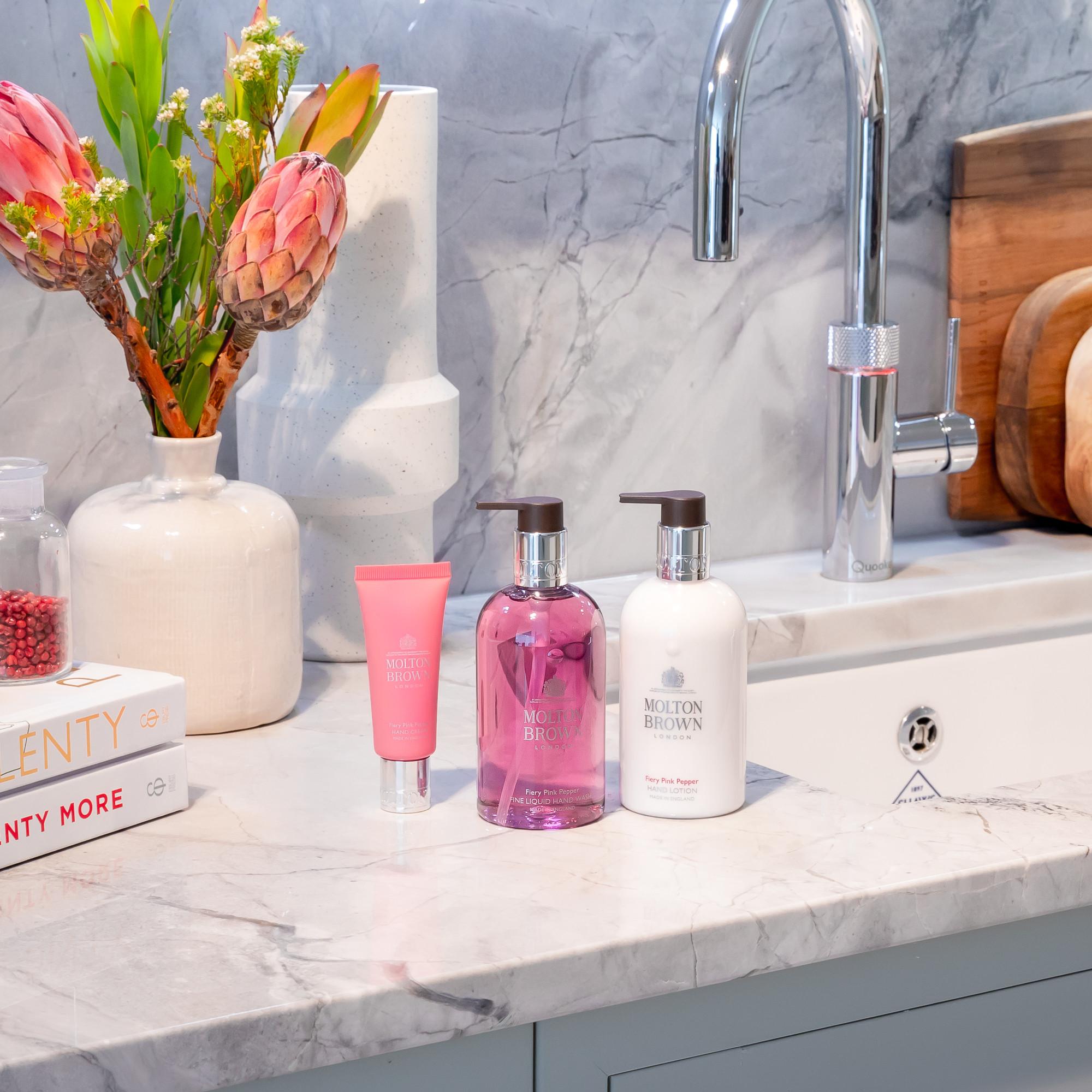 A bathroom sink with silver faucet, MoltonBrown Nederlands Fiery Pink Pepper Handcrème 40ml, white Molton Brown soap bottles, books, a vase of flowers, and wooden cutting boards on a marble countertop.
