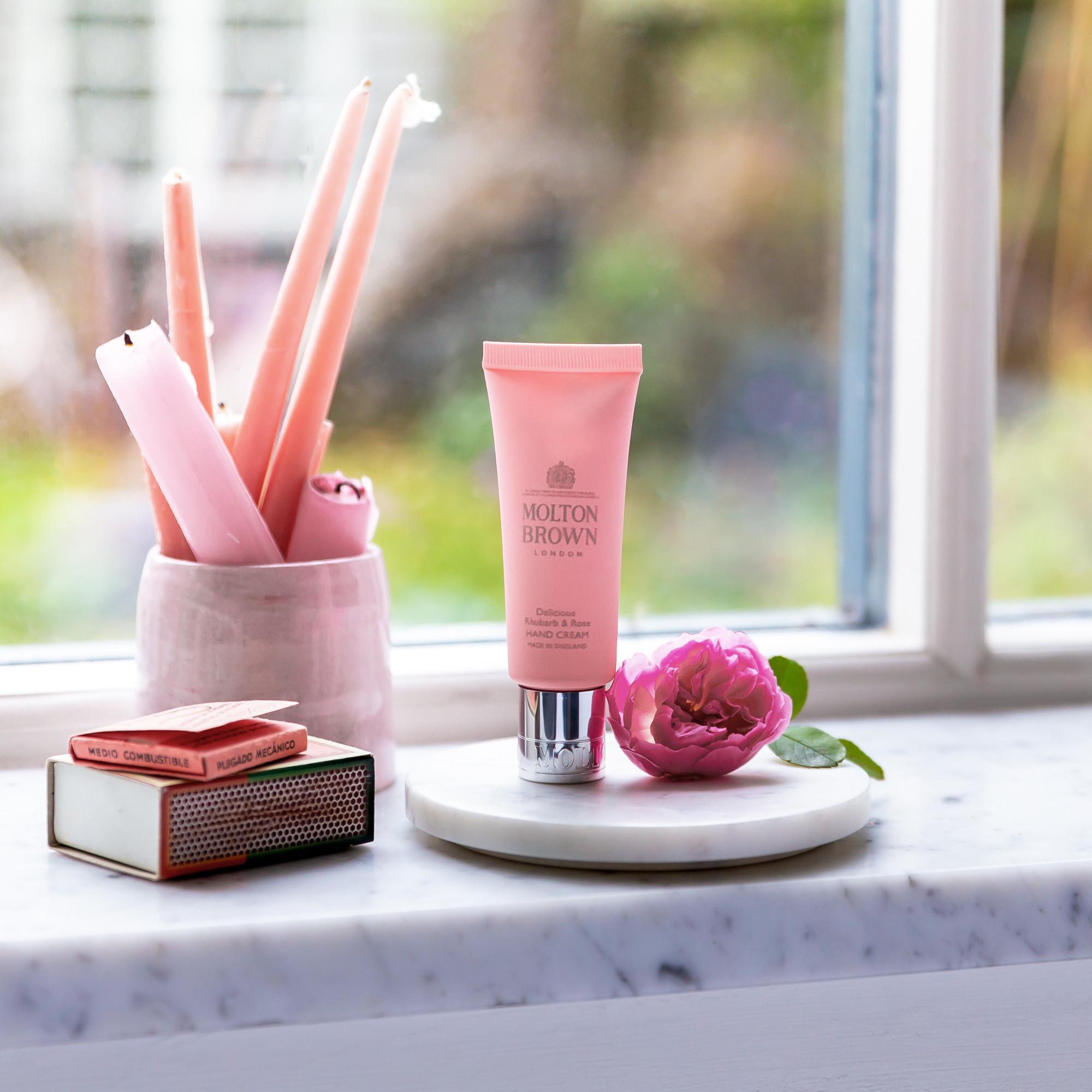 A 40ml tube of MoltonBrown Nederlands Delicious Rhubarb & Rose Handcrème rests on white marble by a window, beside a pink rose, pink candles, and matchboxes—ideal for soothing droge handen with greenery softly blurred outside.