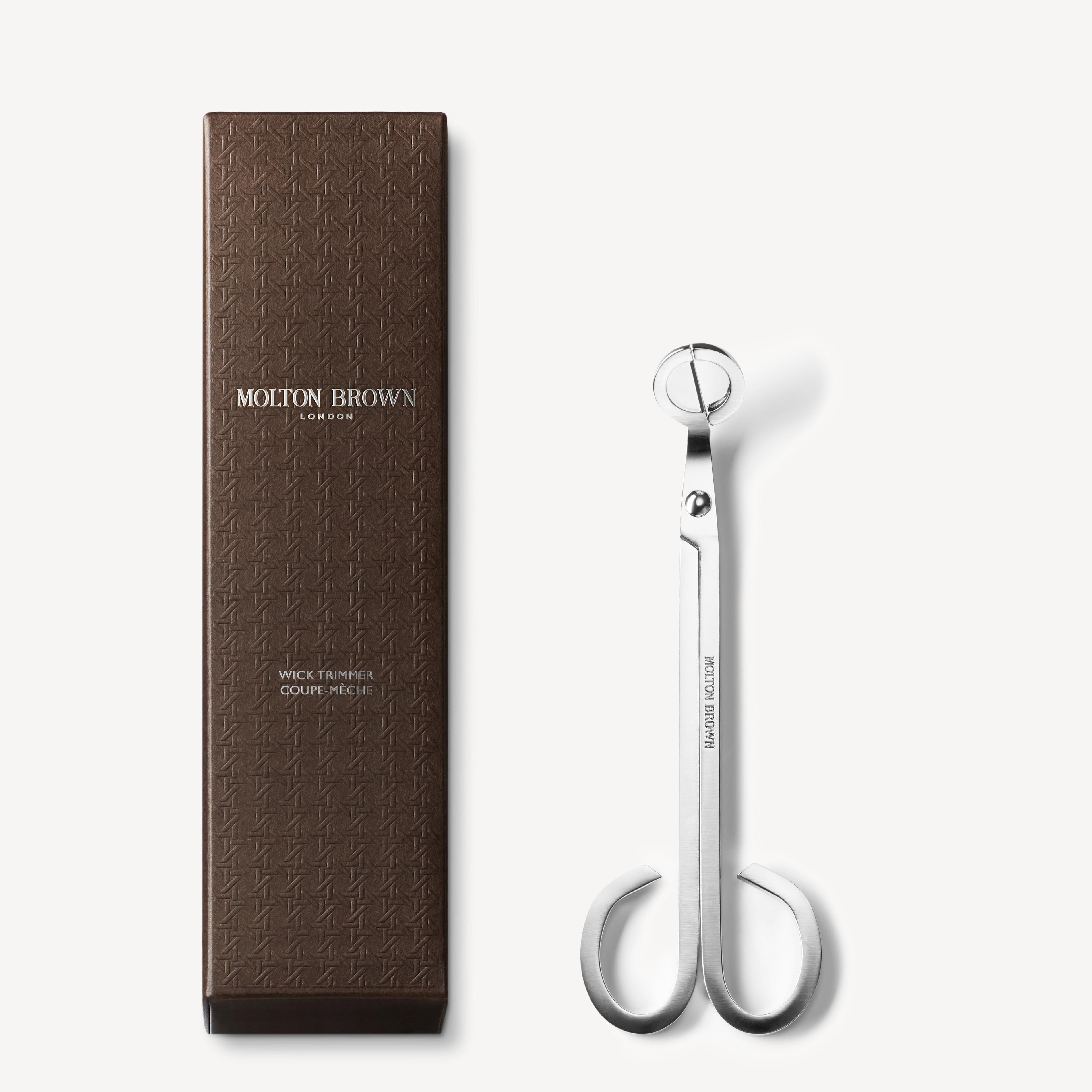 A brown textured box labeled MoltonBrown Nederlands stands next to a sleek, silver Wick Trimmer 72g with rounded handles—an elegant candle accessory—displayed on a white background.